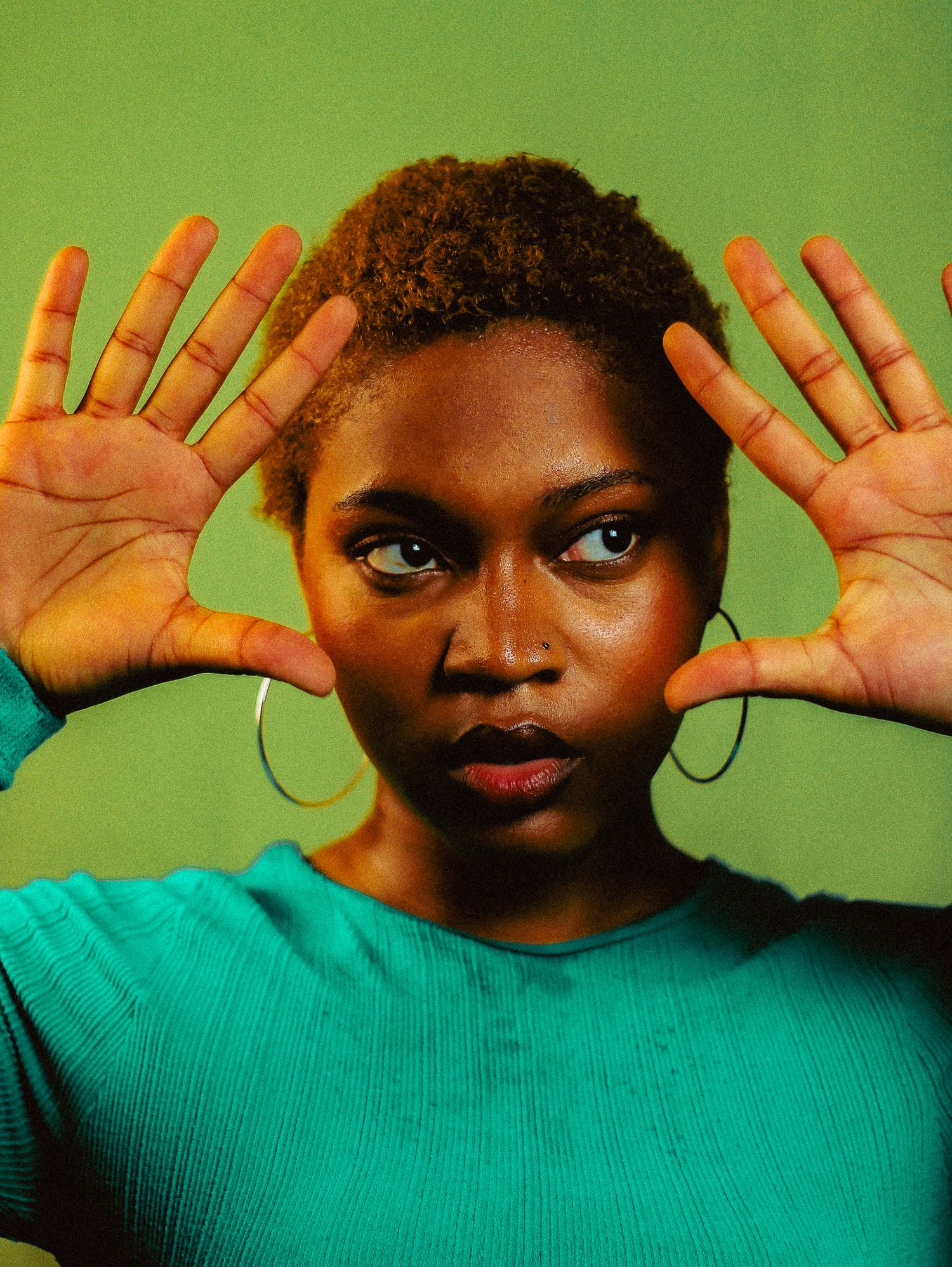 A young Black woman in a teal ribbed sweater raises both hands toward camera on a green background.