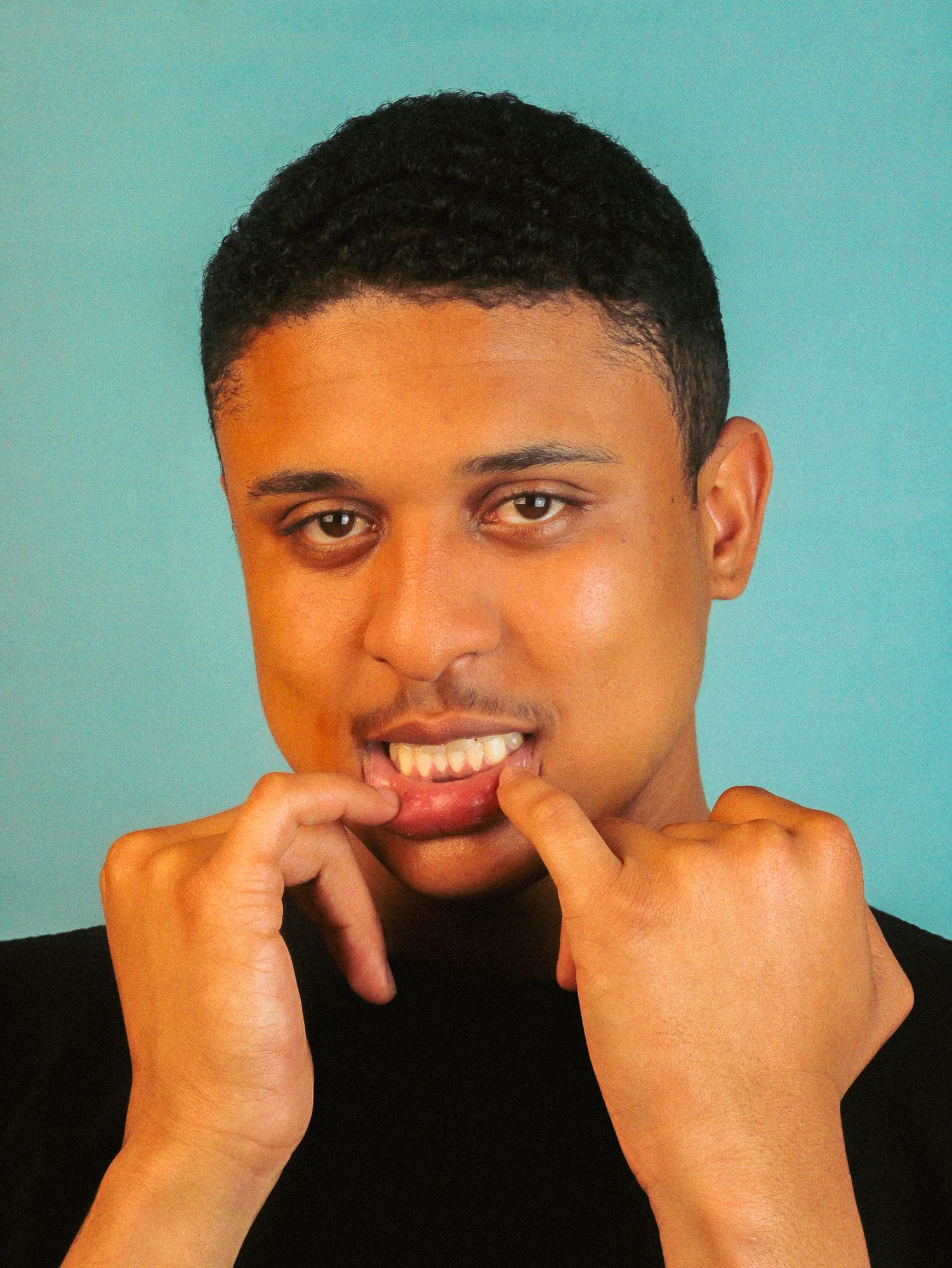 Young man biting his knuckles with an anxious expression against a teal background.