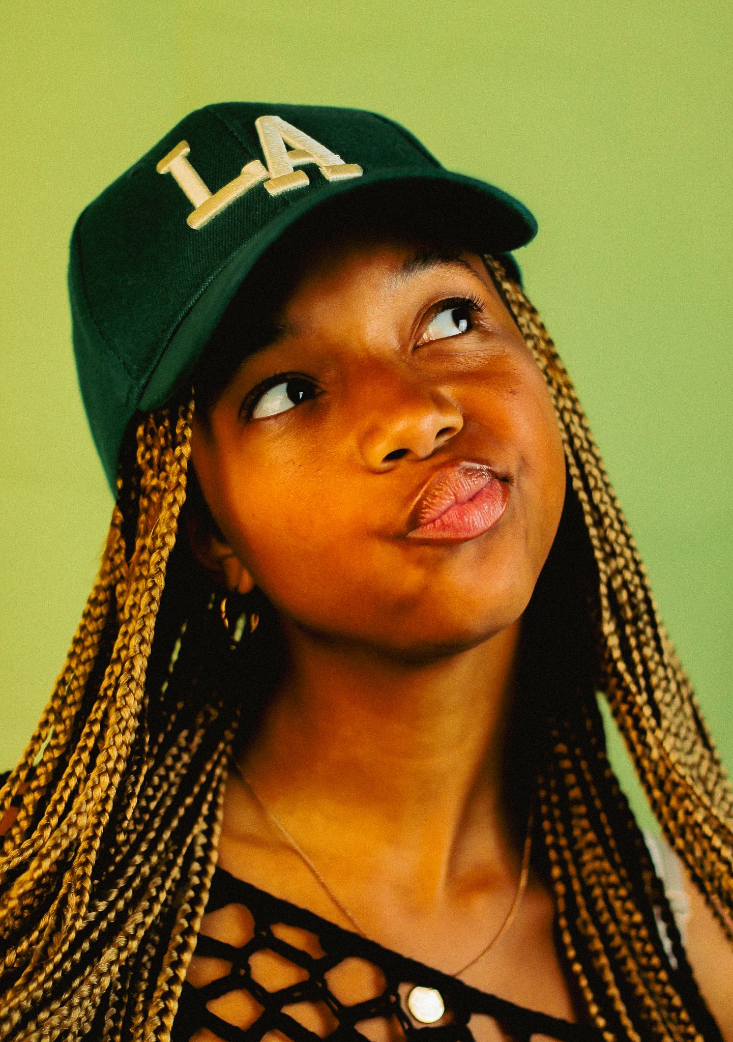 Young Black woman with box braids looking up thoughtfully wearing a green LA snapback cap and hoop earrings.