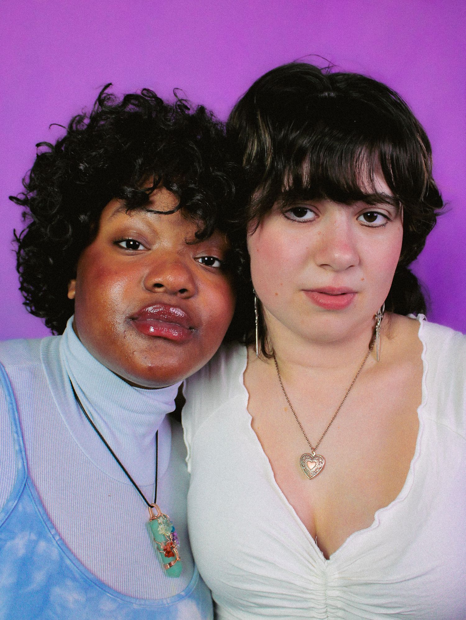 Two young women pose together against a purple background, one in a blue tie-dye top and one in a white top.
