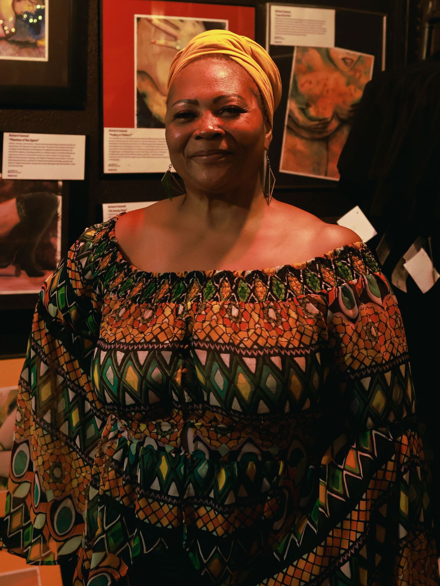 A performer wearing a vibrant African-print dress with geometric patterns stands in front of a gallery wall.