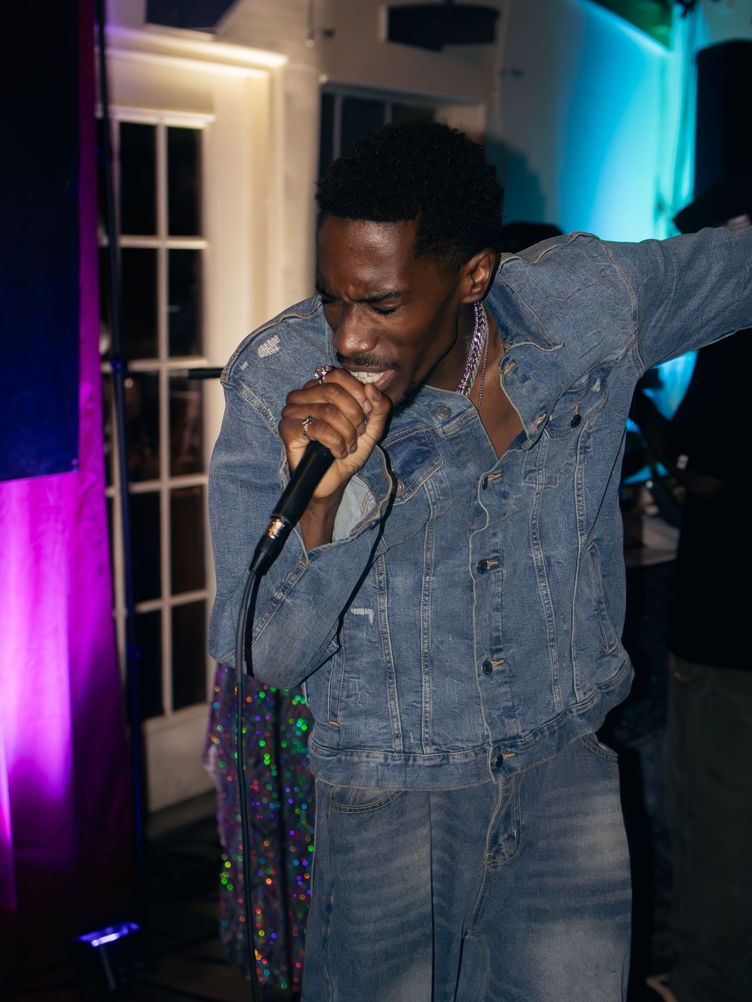Performer grips microphone while wearing full denim outfit under dramatic blue and purple stage lights.