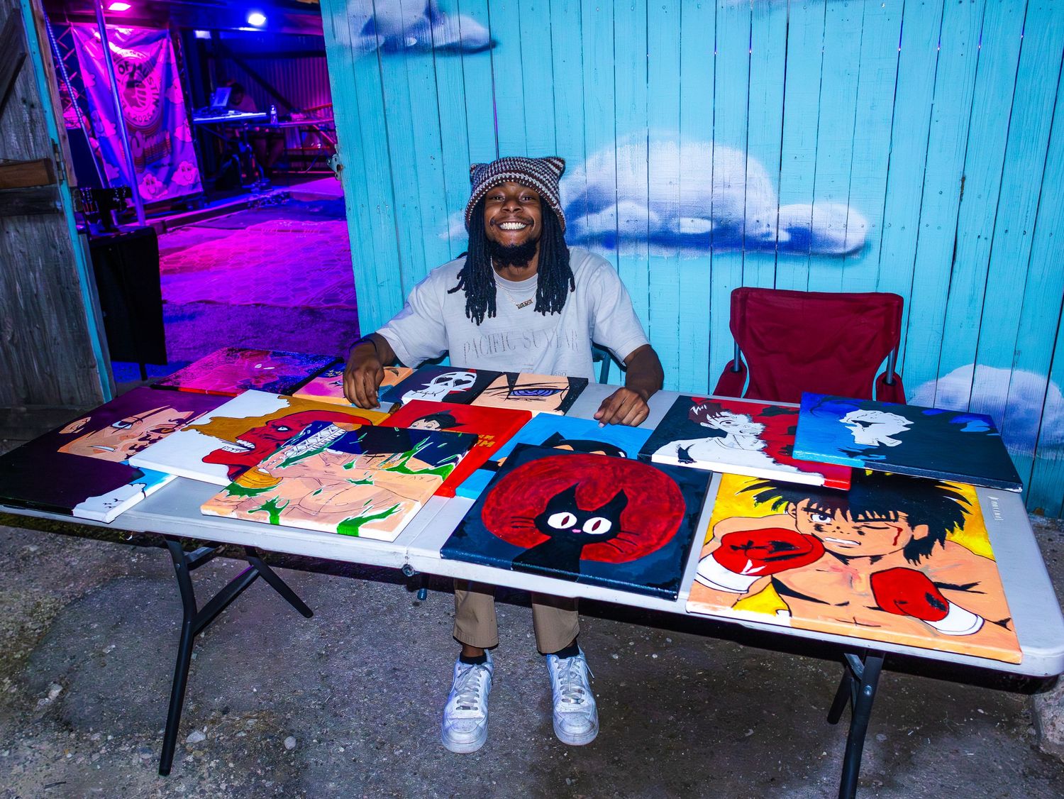 Art booth setup with paintings and prints arranged on tables under colored lighting.