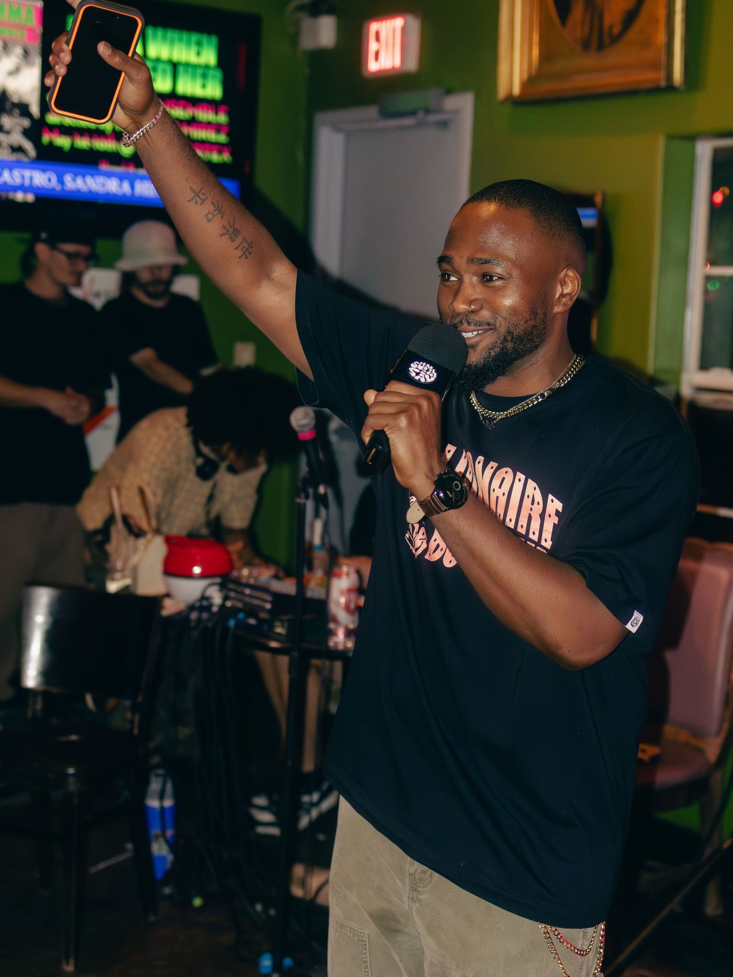 Performer in black t-shirt holding microphone and raising phone during nightclub performance.