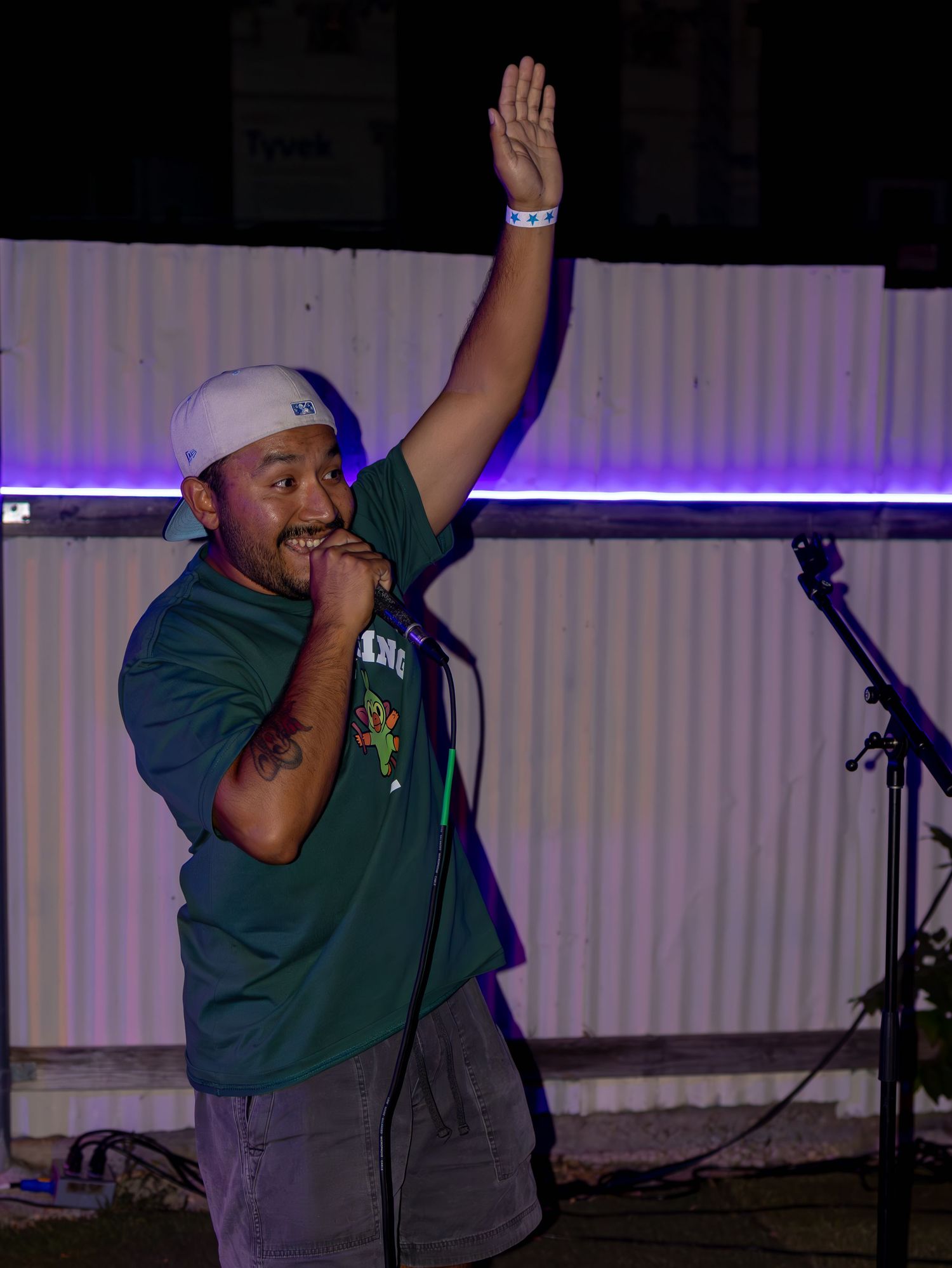 Performer in green t-shirt and baseball cap raises arm during energetic stage performance with purple lighting.