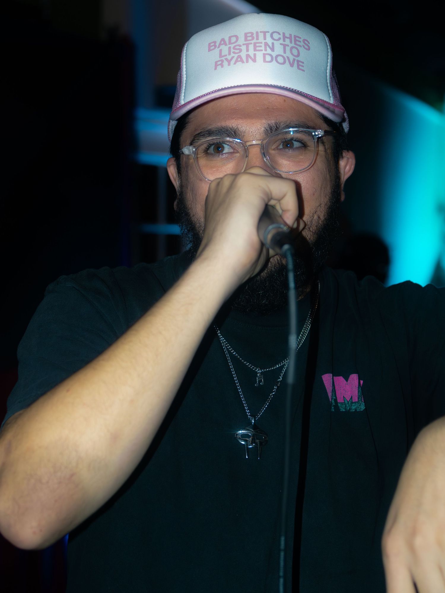 A performer in a baseball cap and glasses holds a microphone while performing under blue lighting.