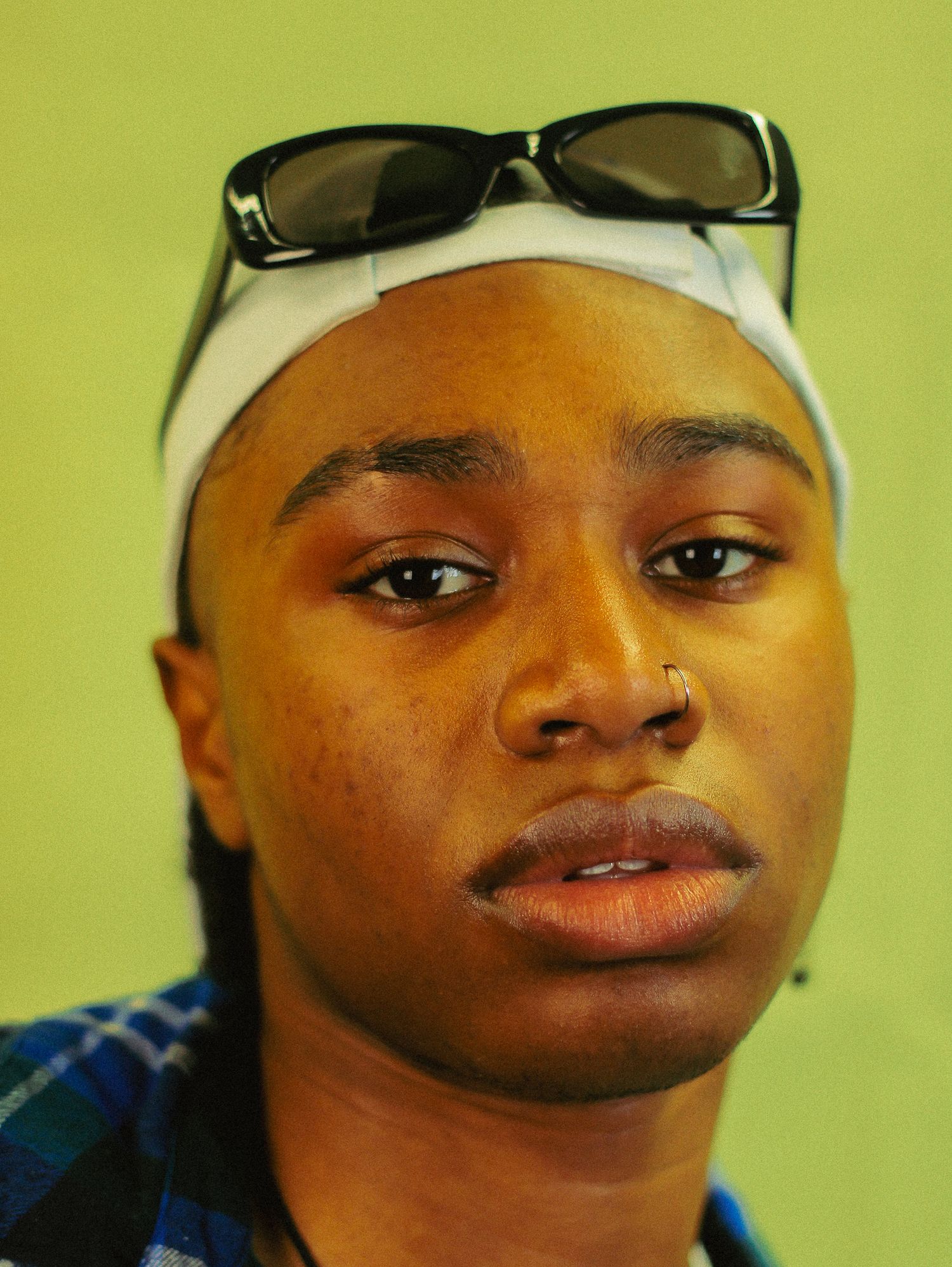 Close-up portrait of young Black woman with white headband and sunglasses on head against green background.