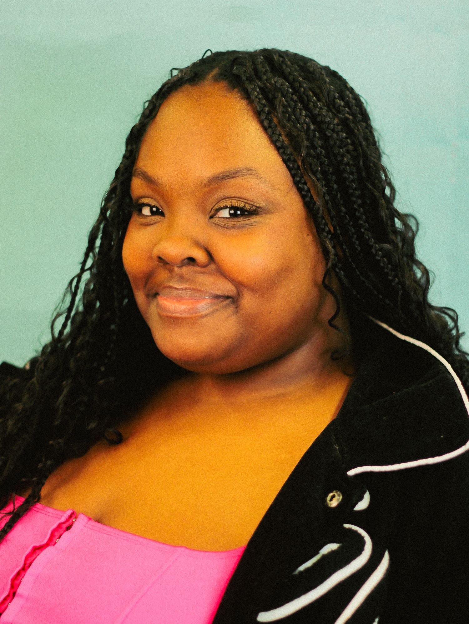 Young woman with long box braids wearing pink top and black jacket smiles against teal background.