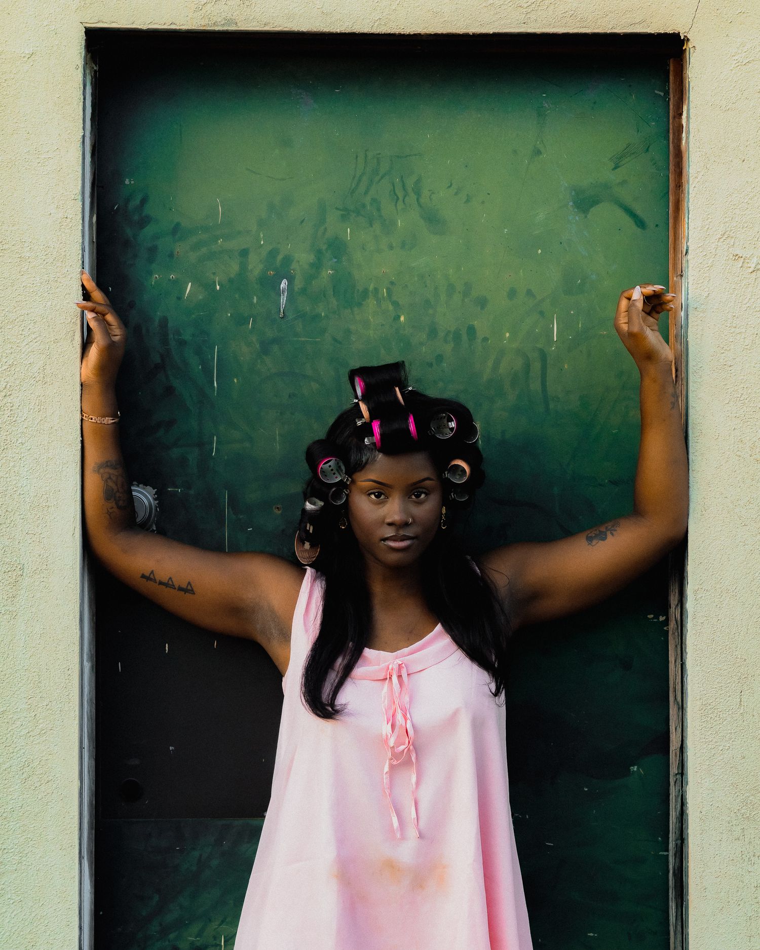 Model in pink dress and curlers poses against green doorway.