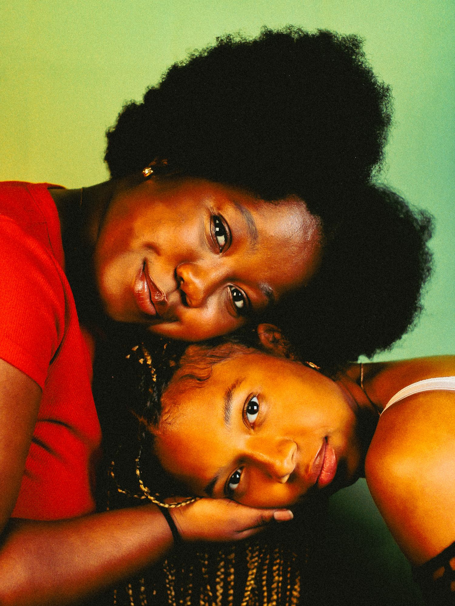 Two Black women posing tenderly together, one with natural afro in red top leaning over the other with braids.