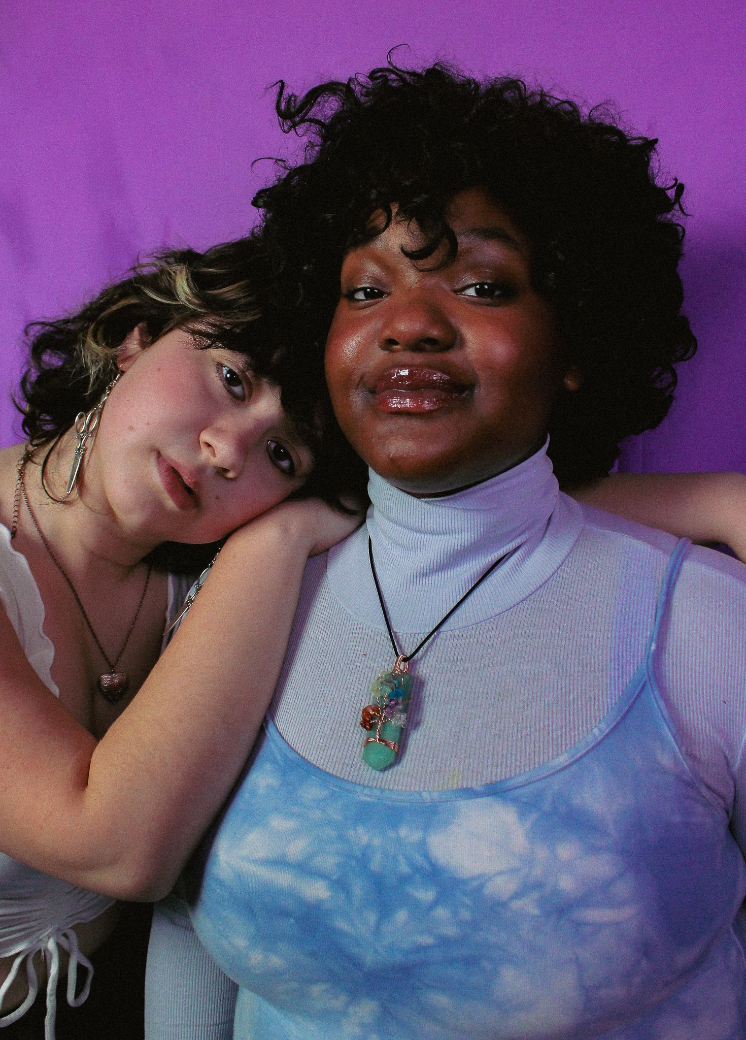 Two young women lean together against a purple background, one resting her head on the other's shoulder.