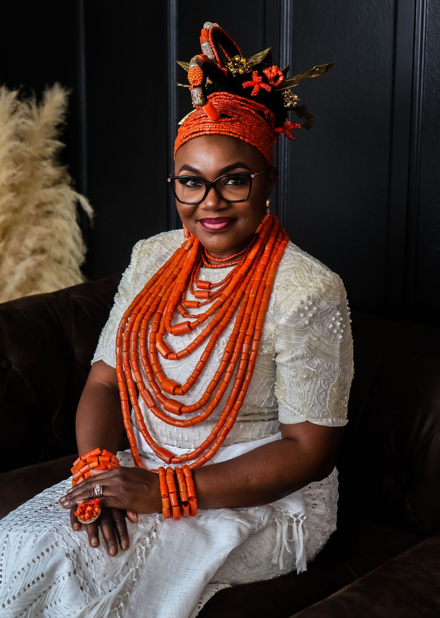 A woman in traditional Nigerian attire with coral beads and an orange headpiece sits elegantly on a dark couch.