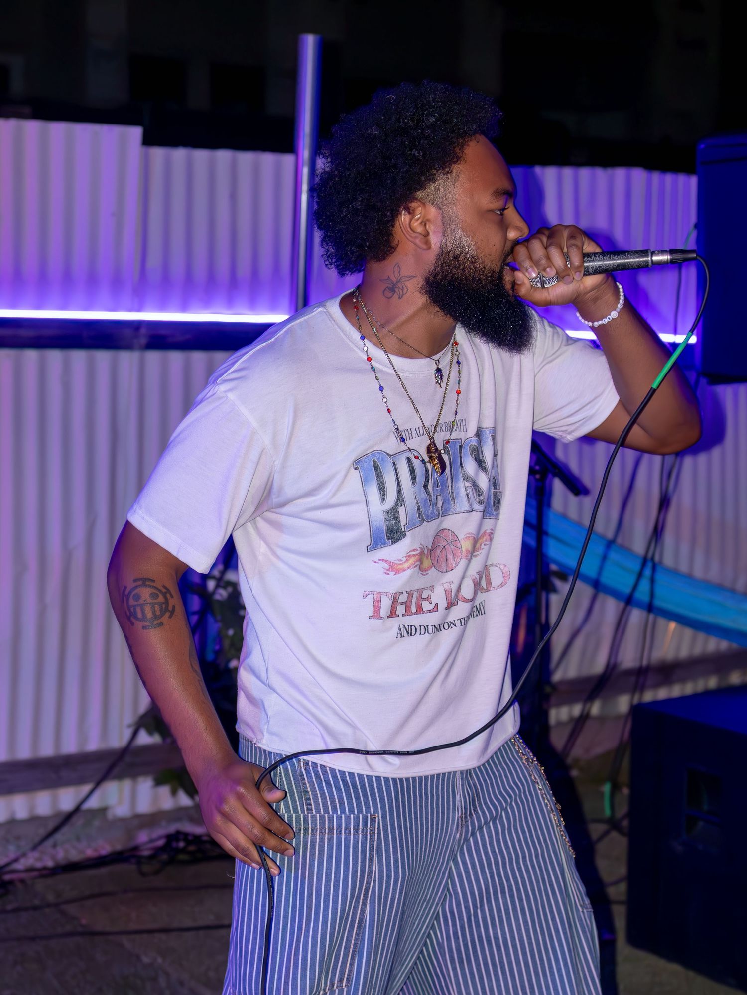 Performer in white tee and striped pants performs with microphone under purple stage lighting.