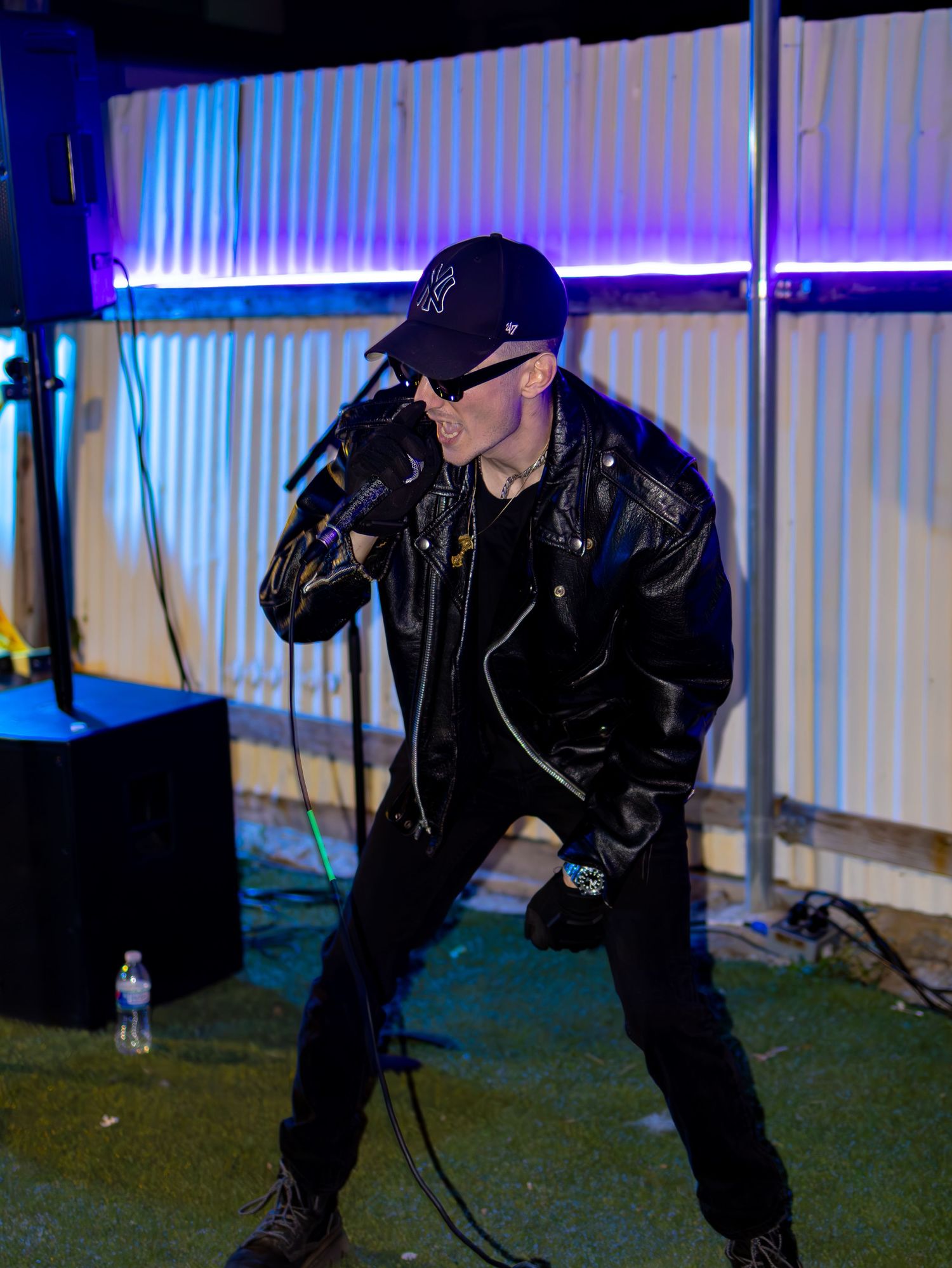 Stage performer in leather jacket strikes pose against colorfully lit background.