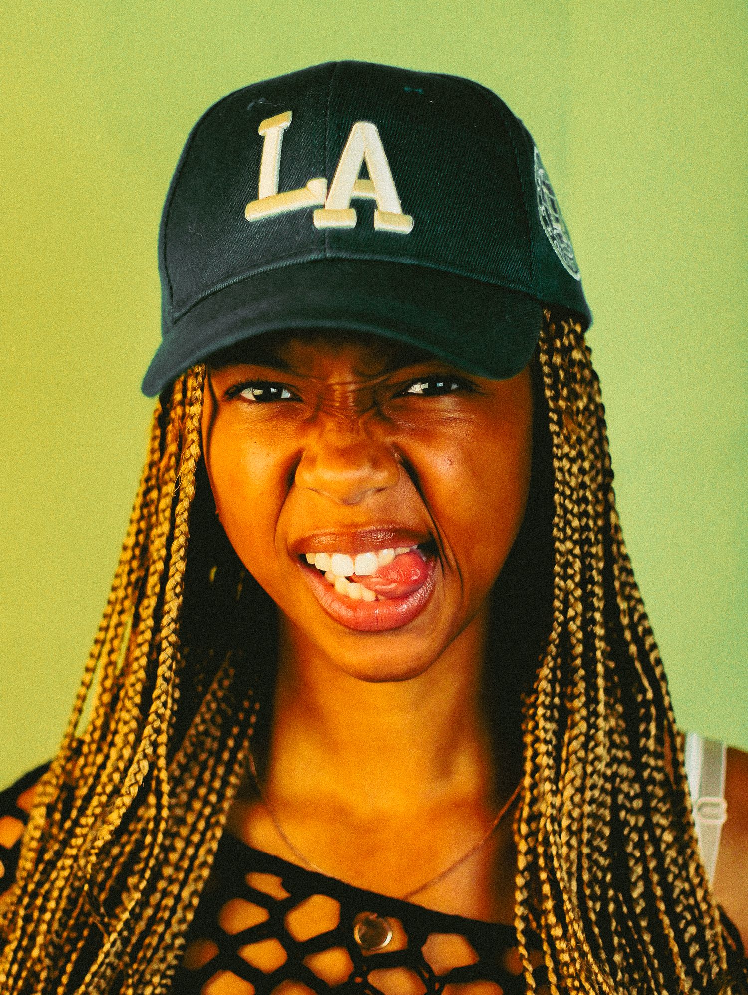 Young woman in LA cap and mesh top makes fierce open-mouth expression against light green background.