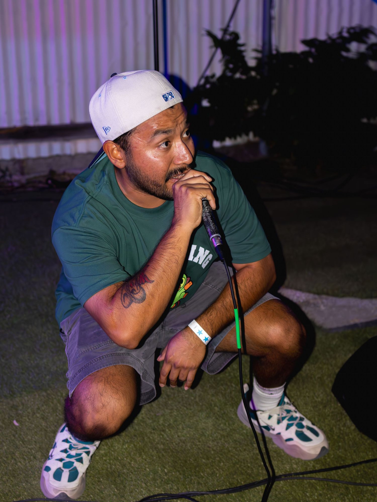 Performer in green shirt and white cap crouches while holding microphone during outdoor performance.