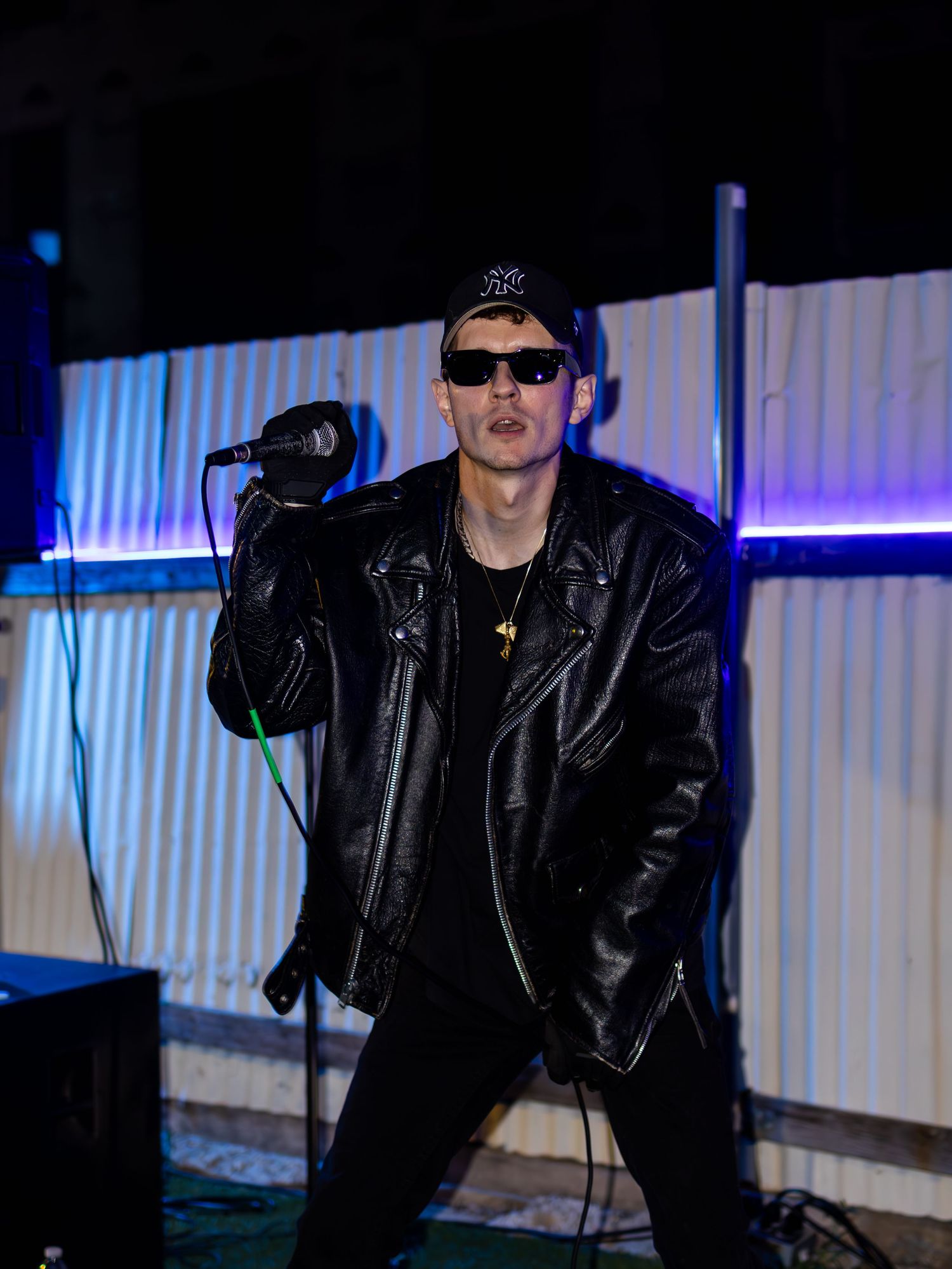 Artist in black leather and baseball cap performs under dramatic stage lighting.