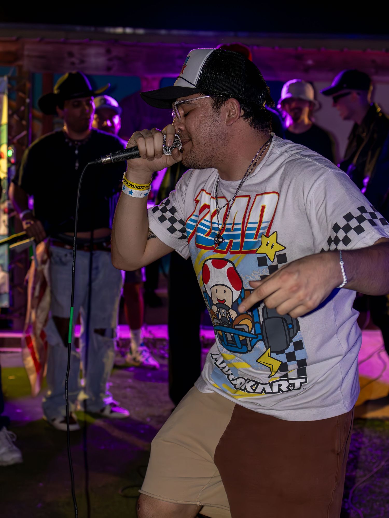 Performer wearing gaming-themed shirt and cap energetically singing into microphone under purple lights.