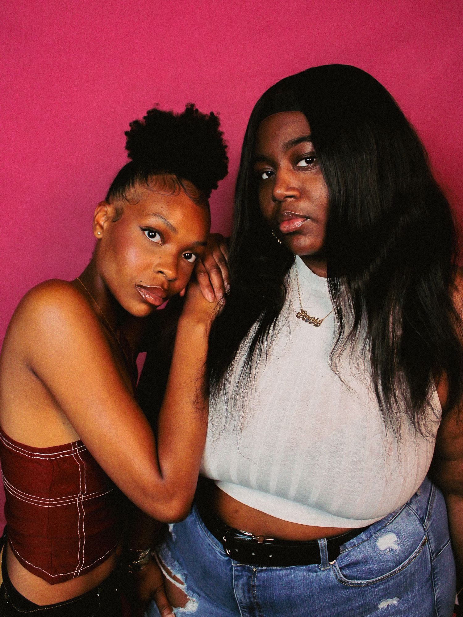 Two friends pose playfully against pink background wearing casual outfits.