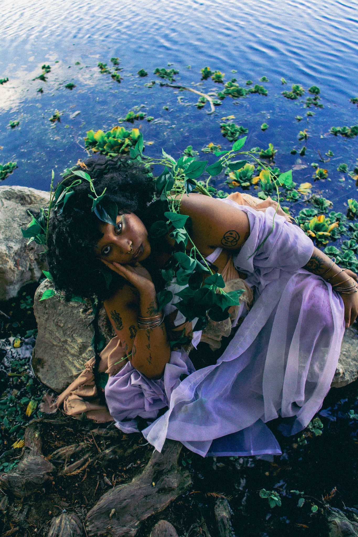 Artistic waterside portrait with flowing lavender fabric and natural elements.