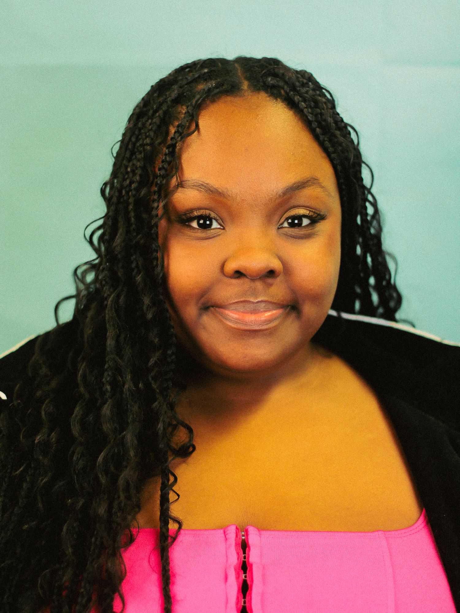 Young woman with long box braids wearing pink strapless top smiles softly against teal background.