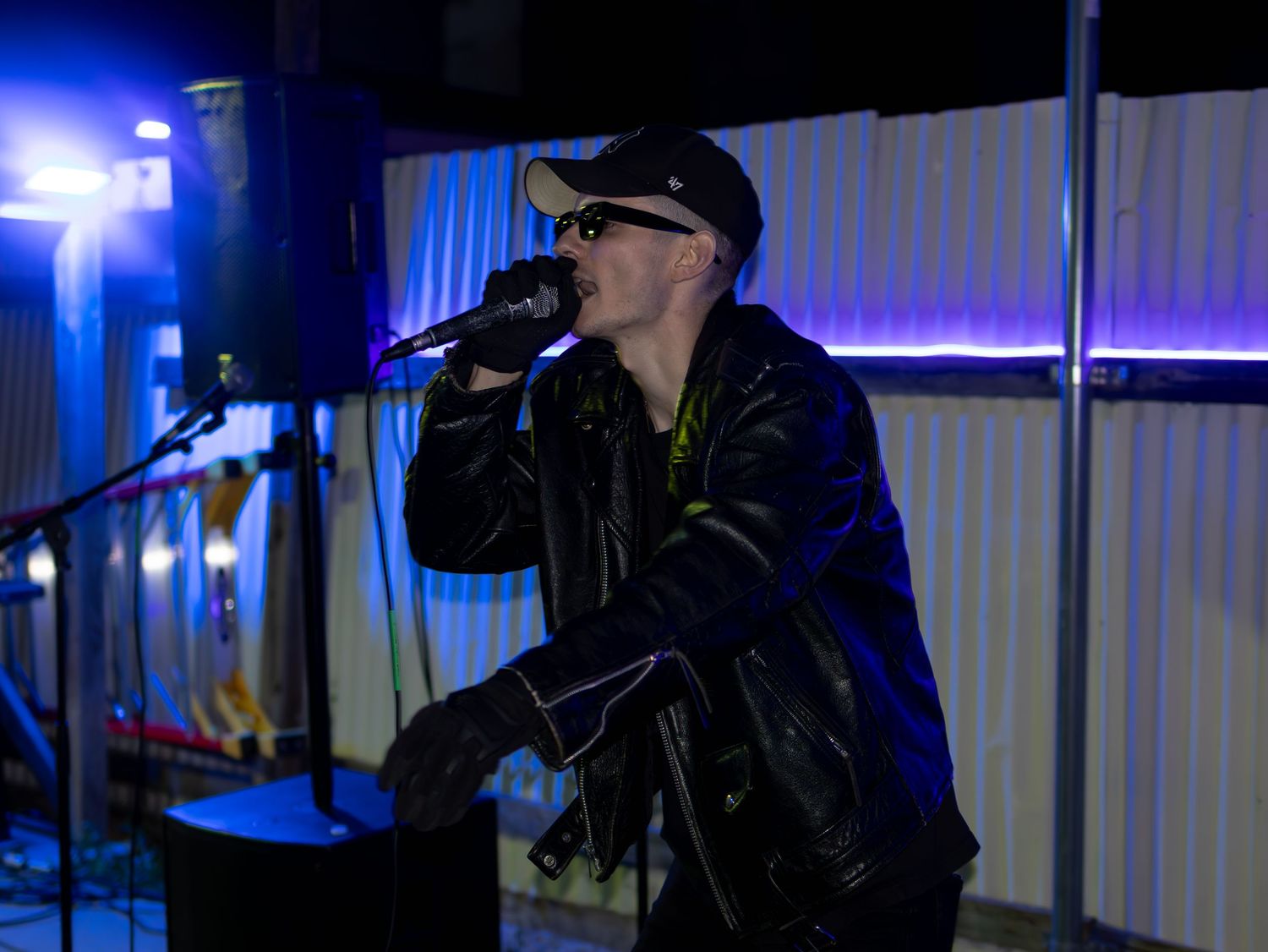 Performer in black leather jacket and baseball cap singing into microphone against blue-lit backdrop.