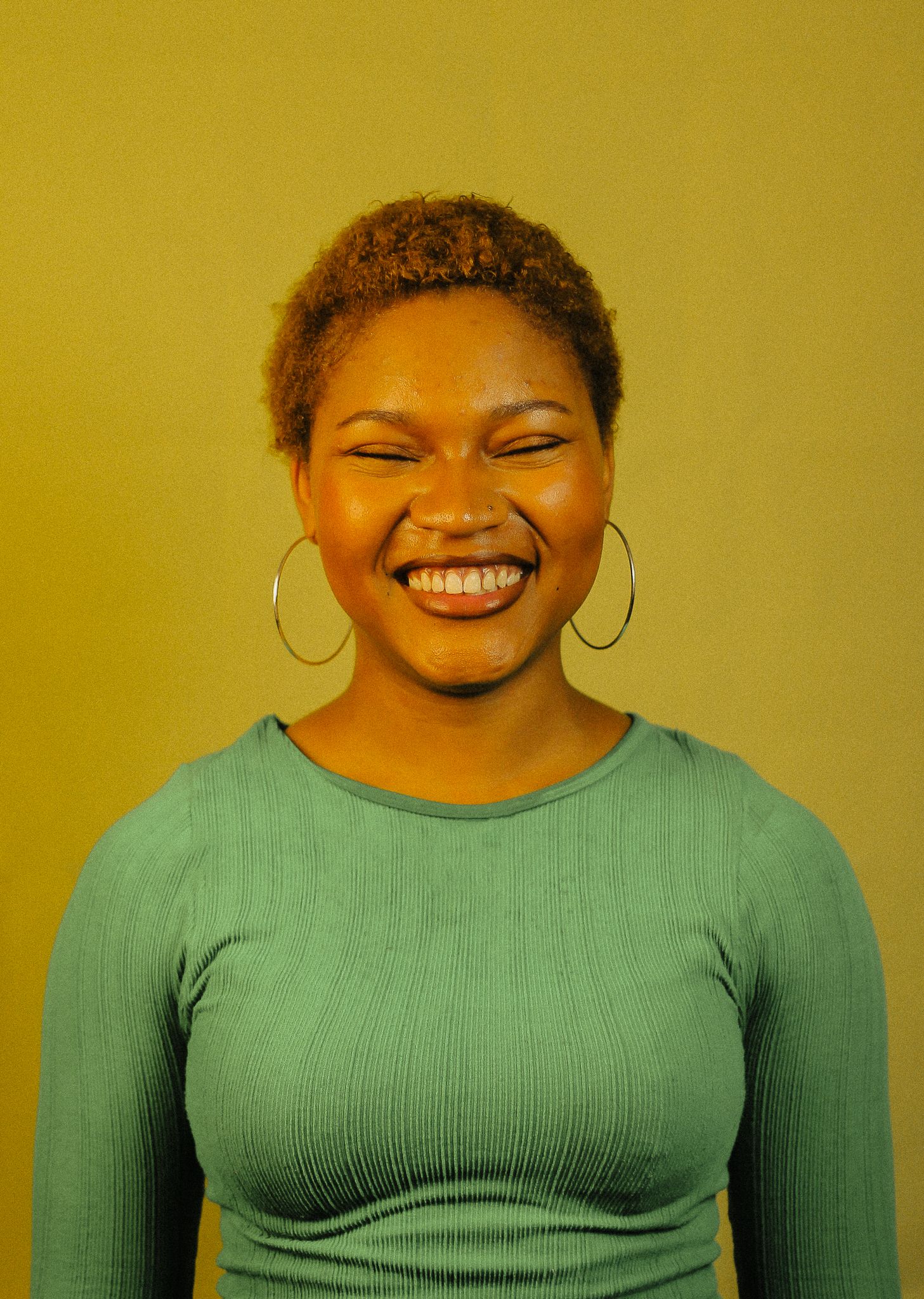 Young woman with short natural hair laughing with eyes closed, wearing teal ribbed top and hoop earrings.