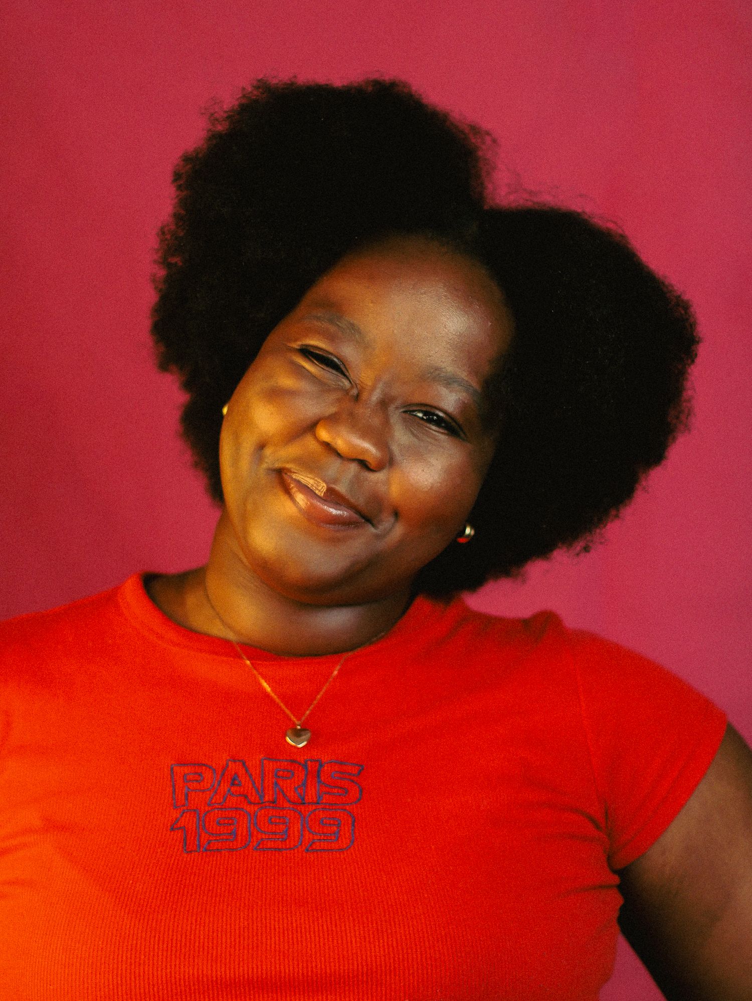Smiling Black woman with natural afro hair wearing a red Paris 1999 t-shirt against a pink background.