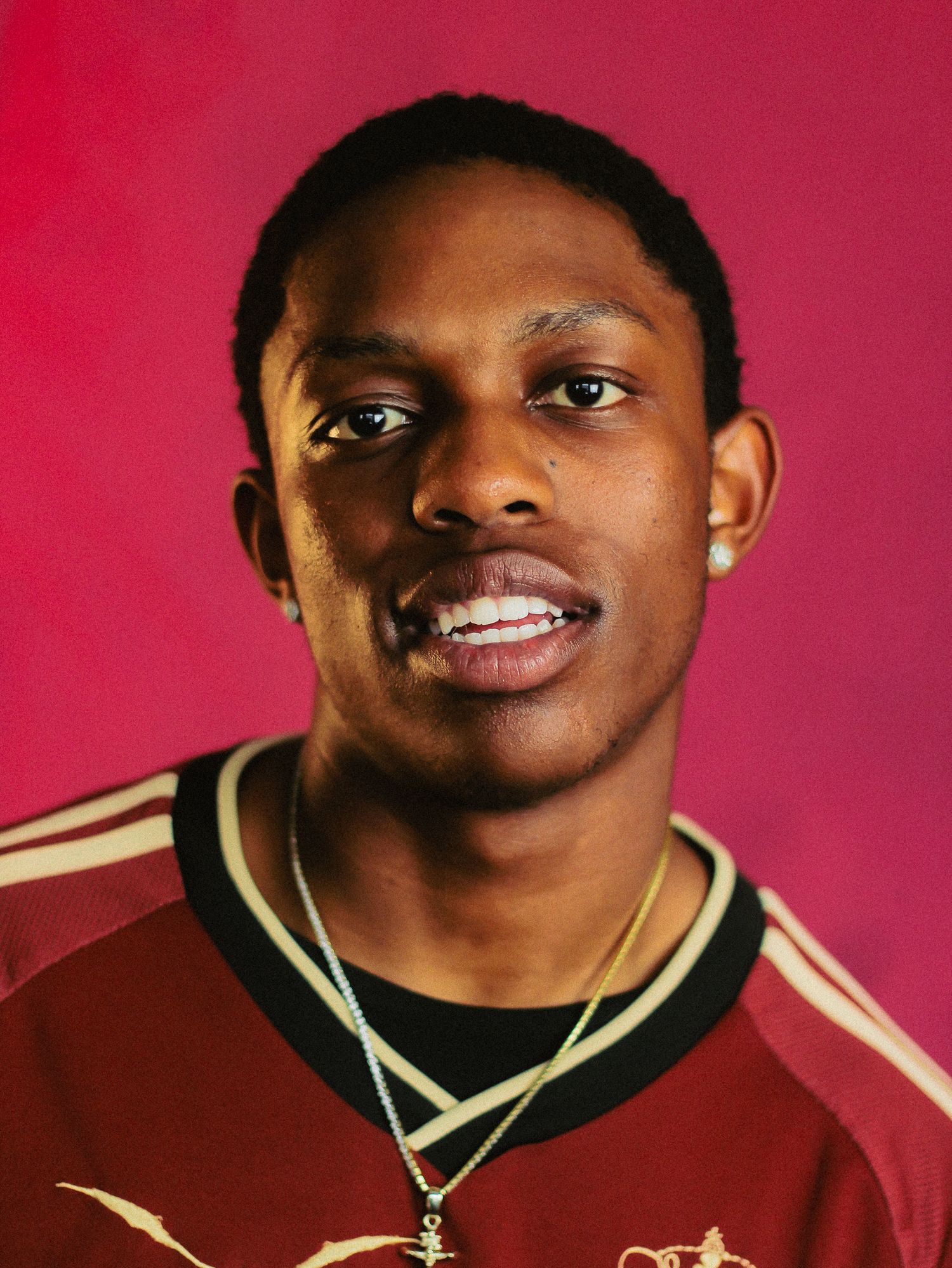Young Black man in maroon soccer jersey smiles wearing gold chain against pink background.