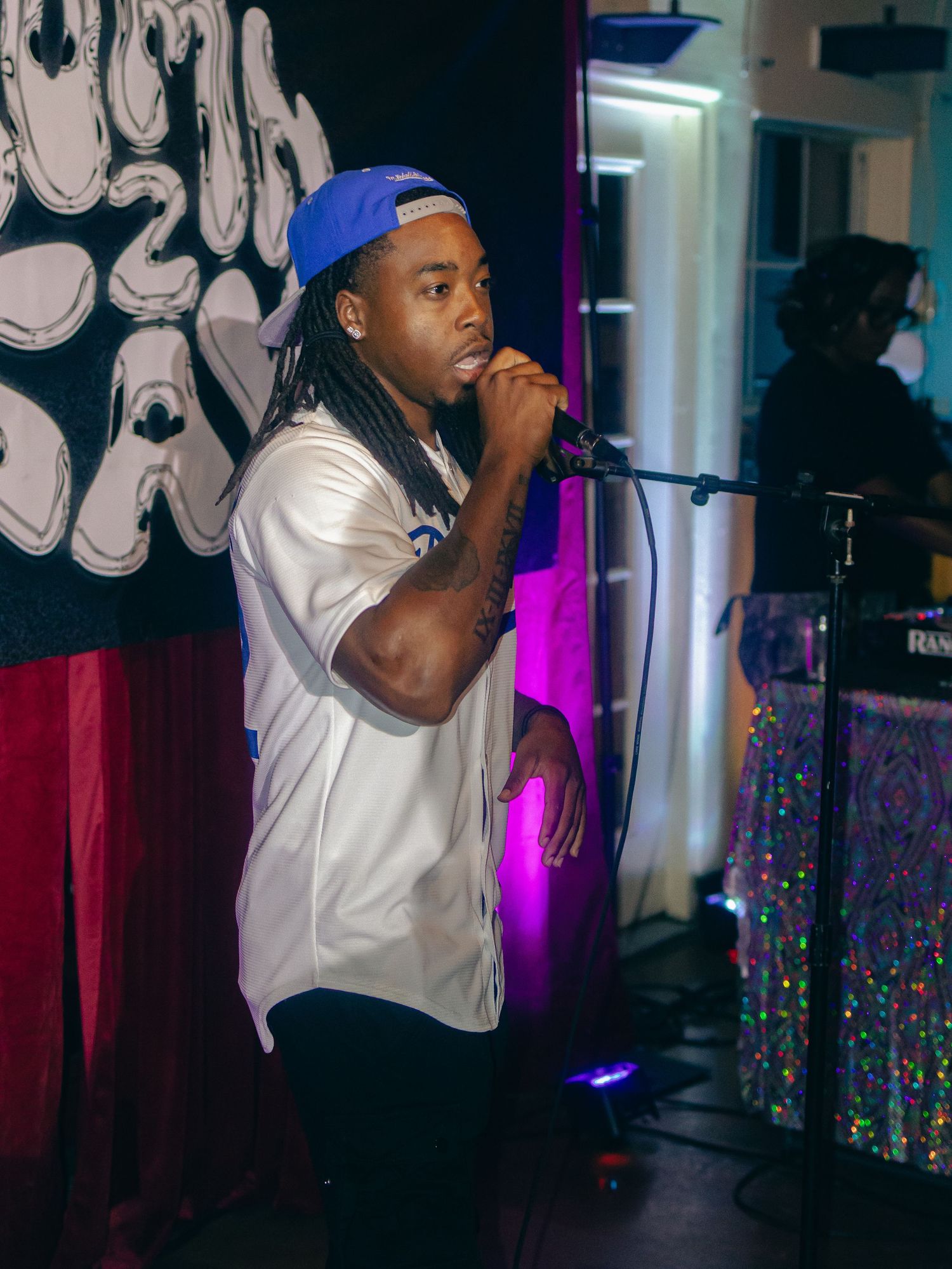 Artist in baseball cap and white shirt performing against graffiti-style wall backdrop.