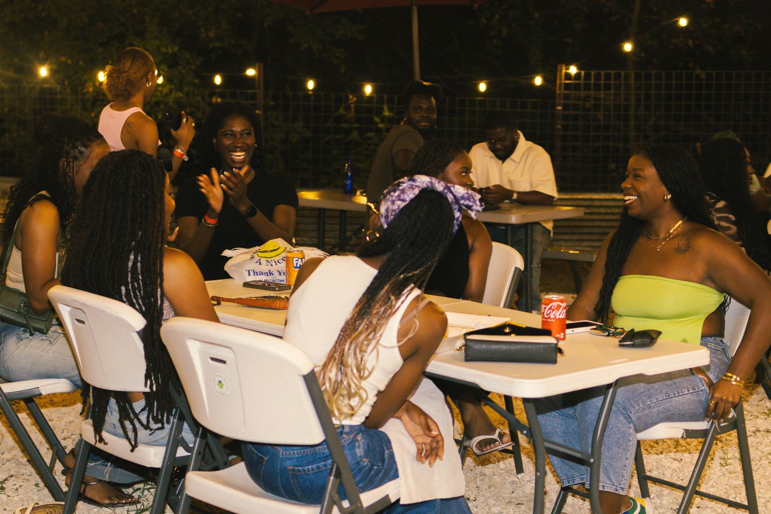 People gathered at outdoor tables for a nighttime social event under string lights.