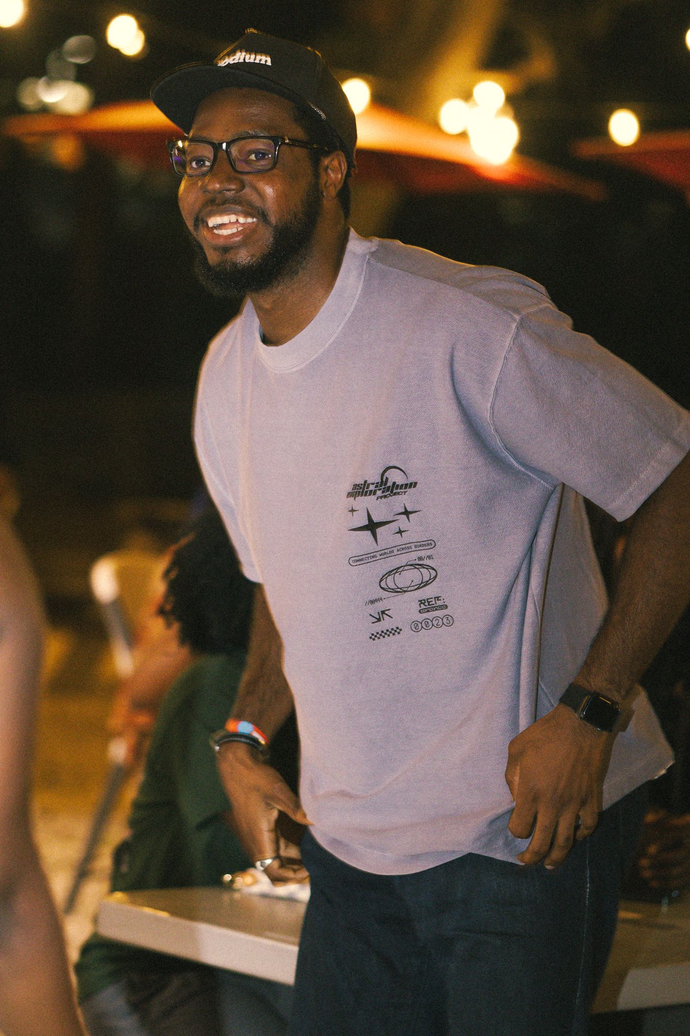 Person wearing a light purple t-shirt with graphics smiling in dim lighting.