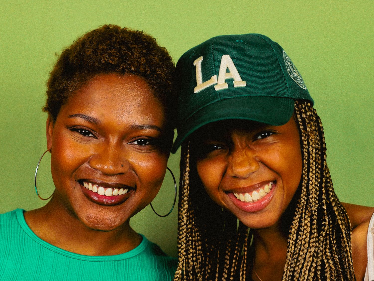 Two smiling women pose together, one in teal top and one wearing an LA baseball cap against green background.