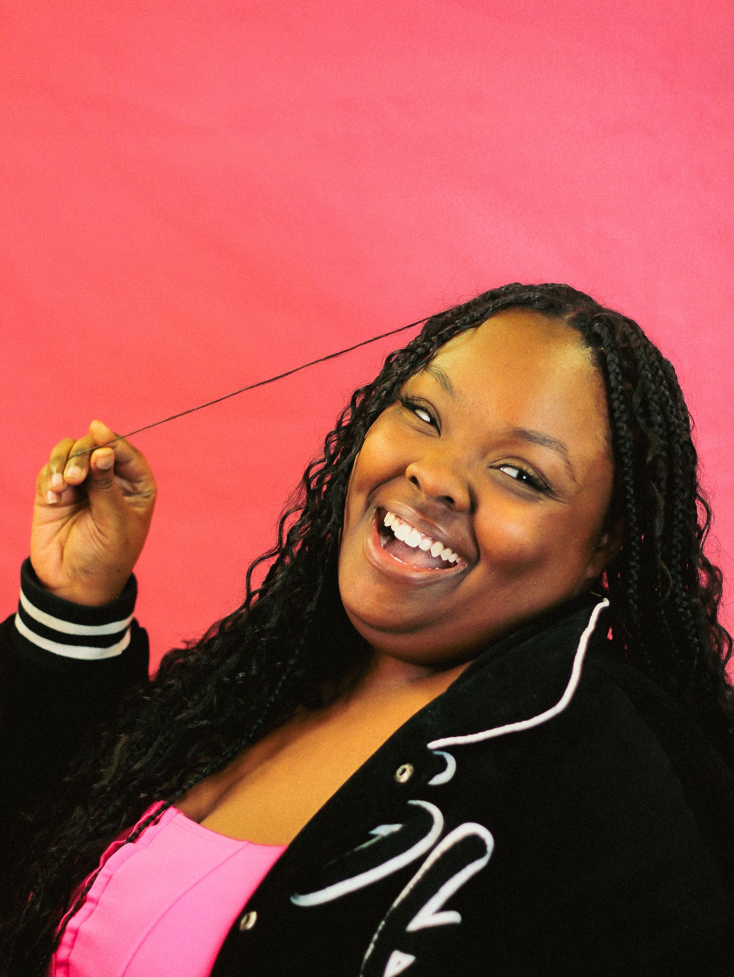Woman with long braids laughs while pulling a strand of hair, wearing a black jacket against a pink background.