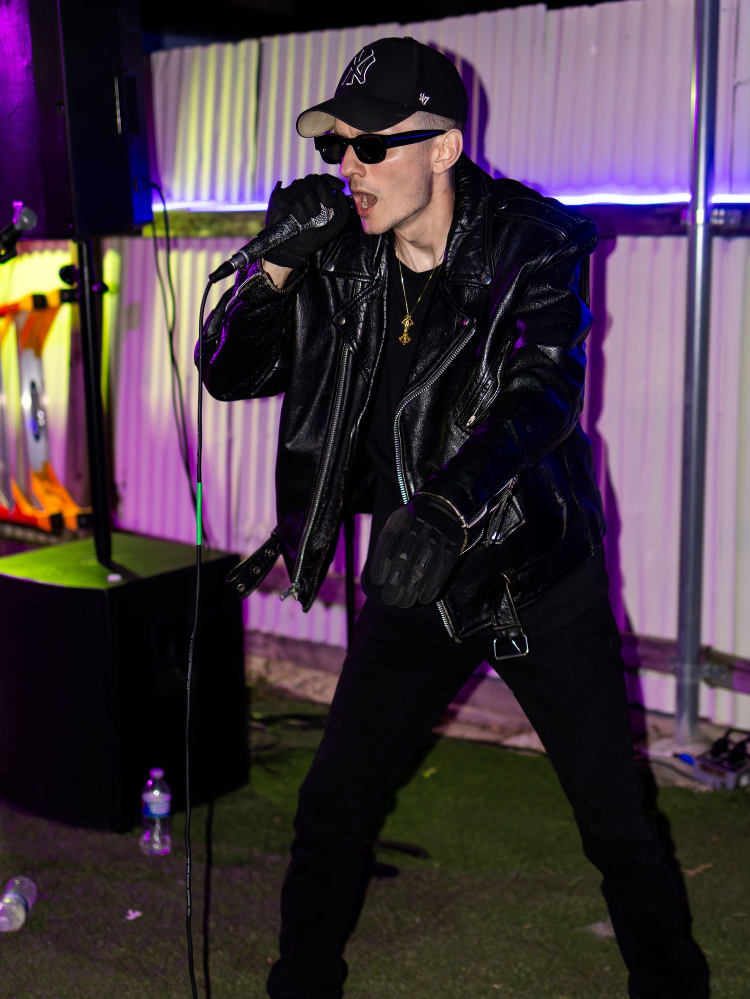 Performer in black leather jacket and sunglasses strikes dynamic pose on stage with colorful lighting.