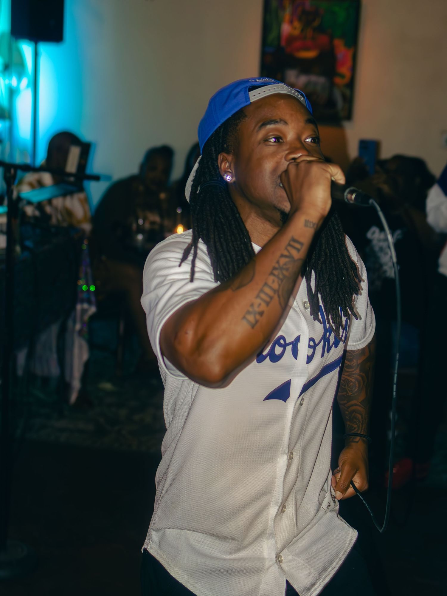 Performer in white baseball jersey and blue cap singing with tattoos visible under stage lights.