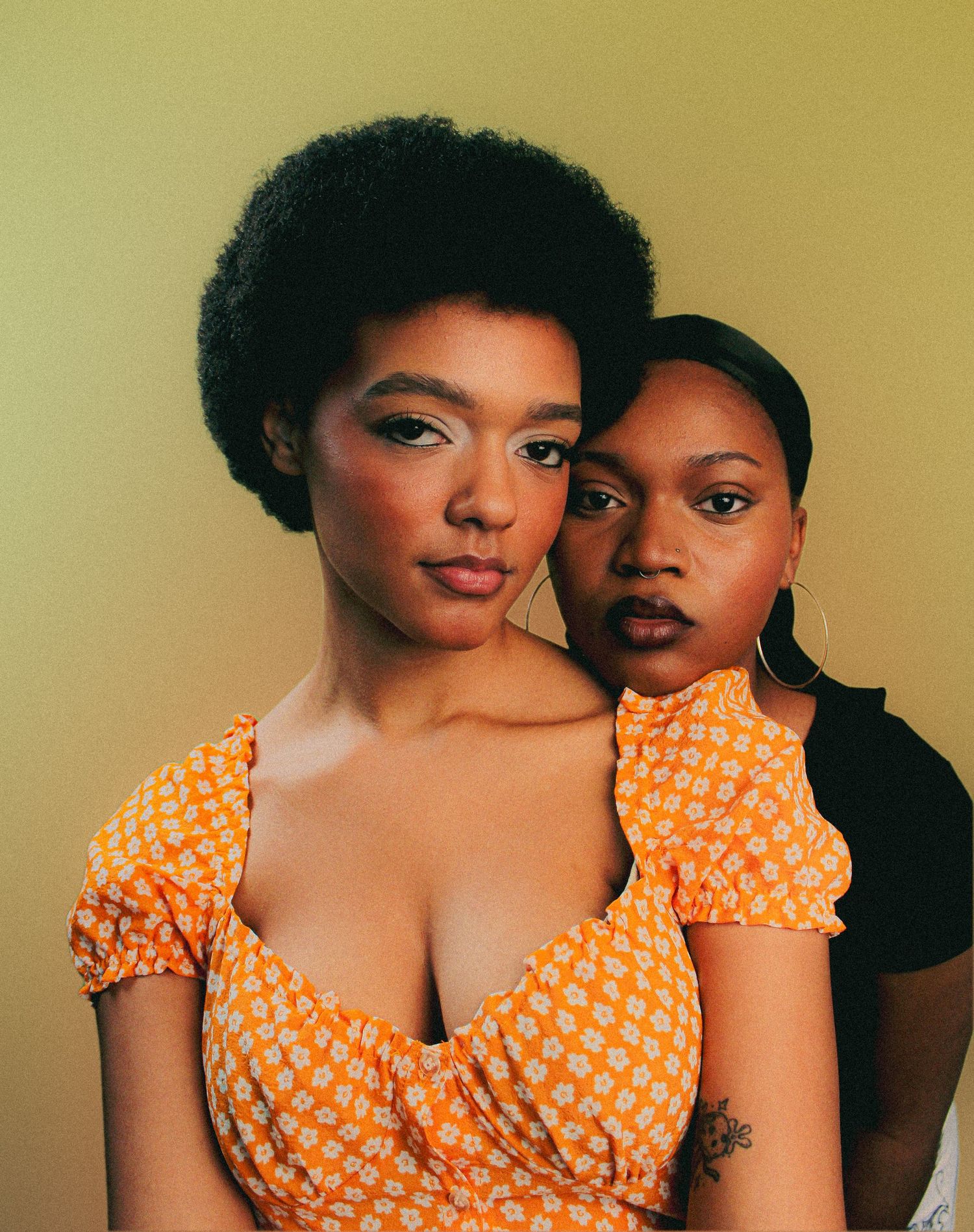 Two people in orange polka dot dress and dark clothing pose intimately against a warm yellow background.