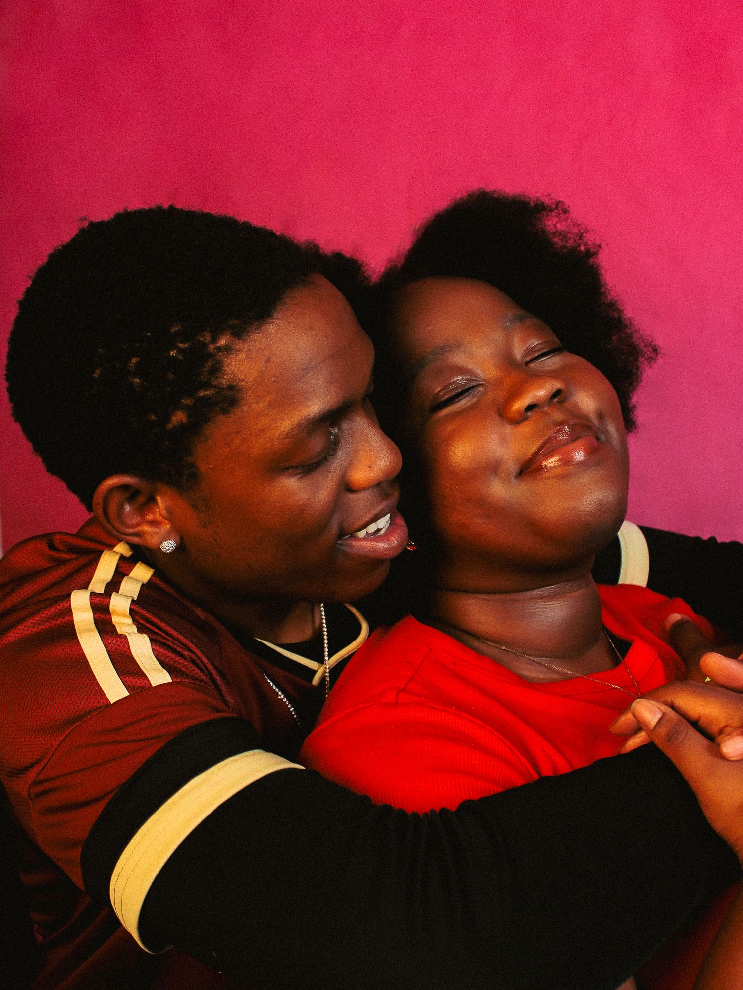 Two people sharing a joyful embrace against a pink background, one hugging the other from behind while both smile.
