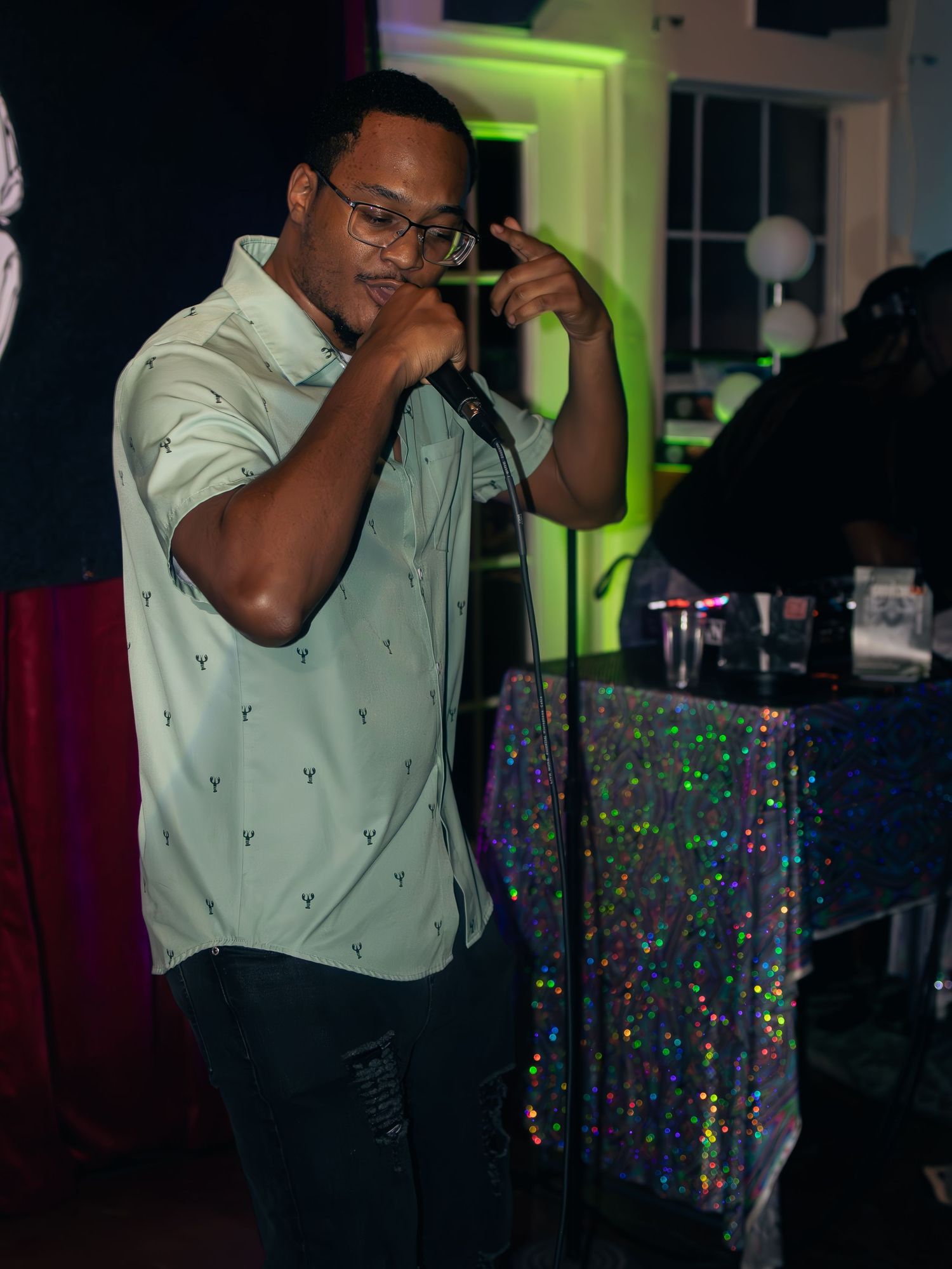 Singer in light green patterned shirt performs energetically with sparkly backdrop visible.