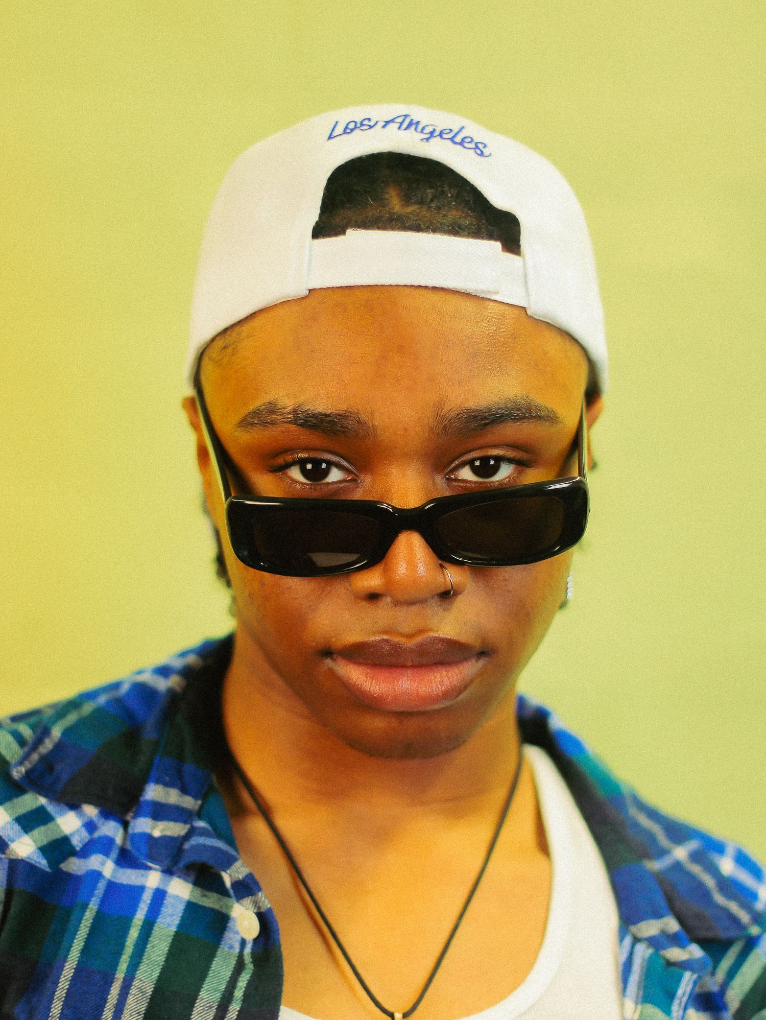Young person wearing white Los Angeles snapback cap and black rectangular sunglasses against green background.
