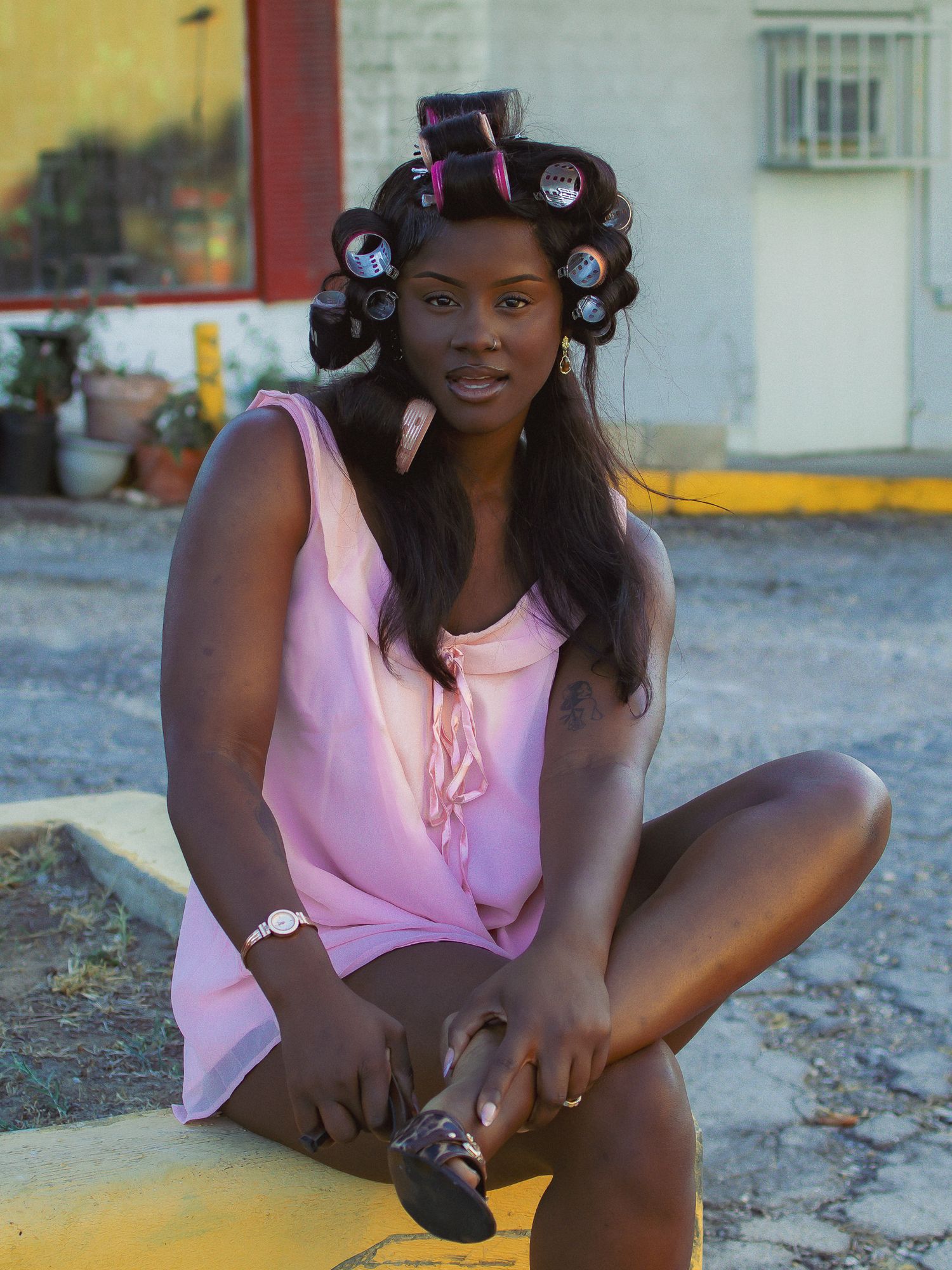 A fashion photoshoot at sunset featuring a pink sleeveless top and hair curlers against a rustic backdrop.