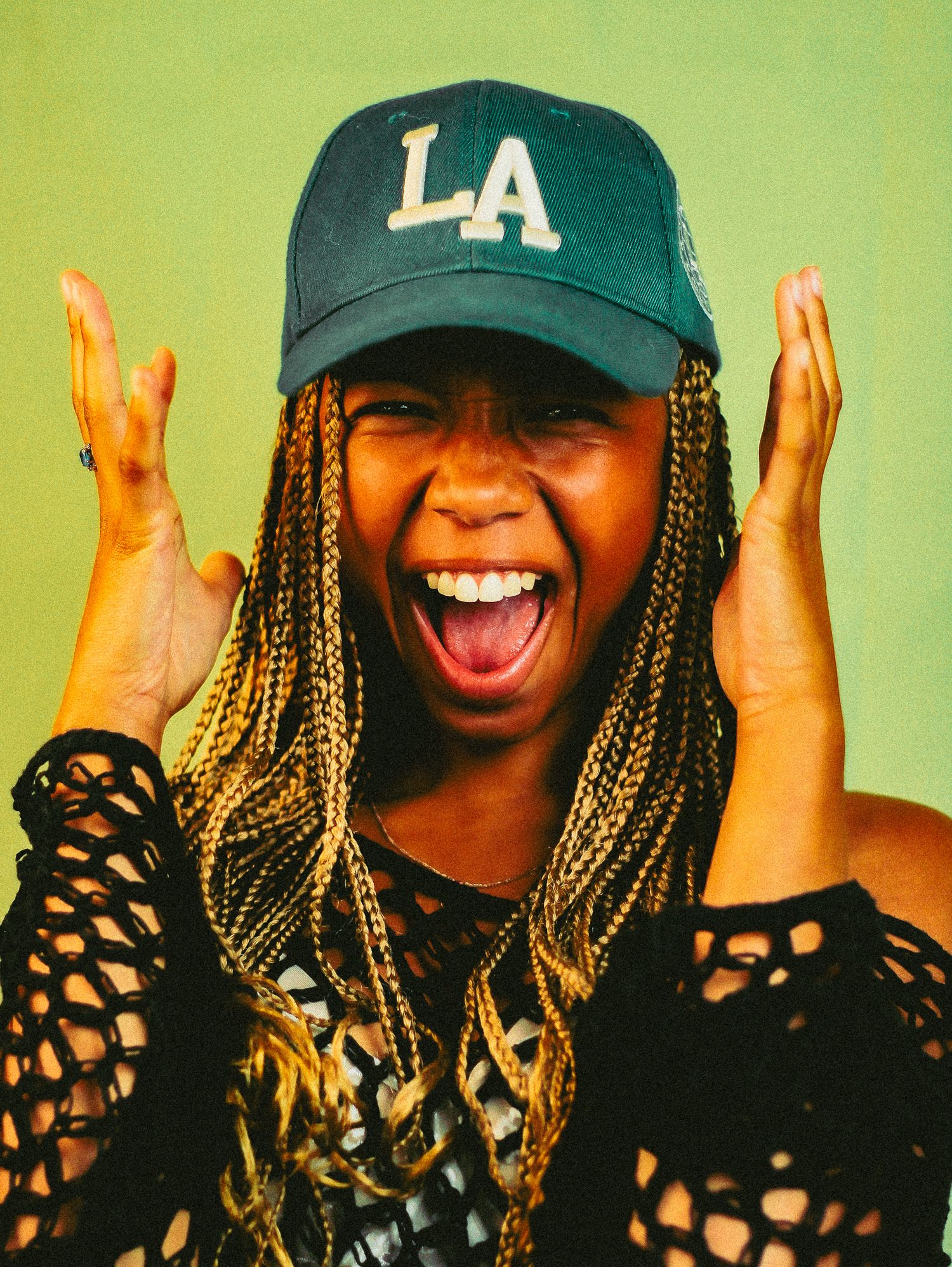 Excited young Black woman with braids laughing with hands raised wearing a navy LA cap against green backdrop.