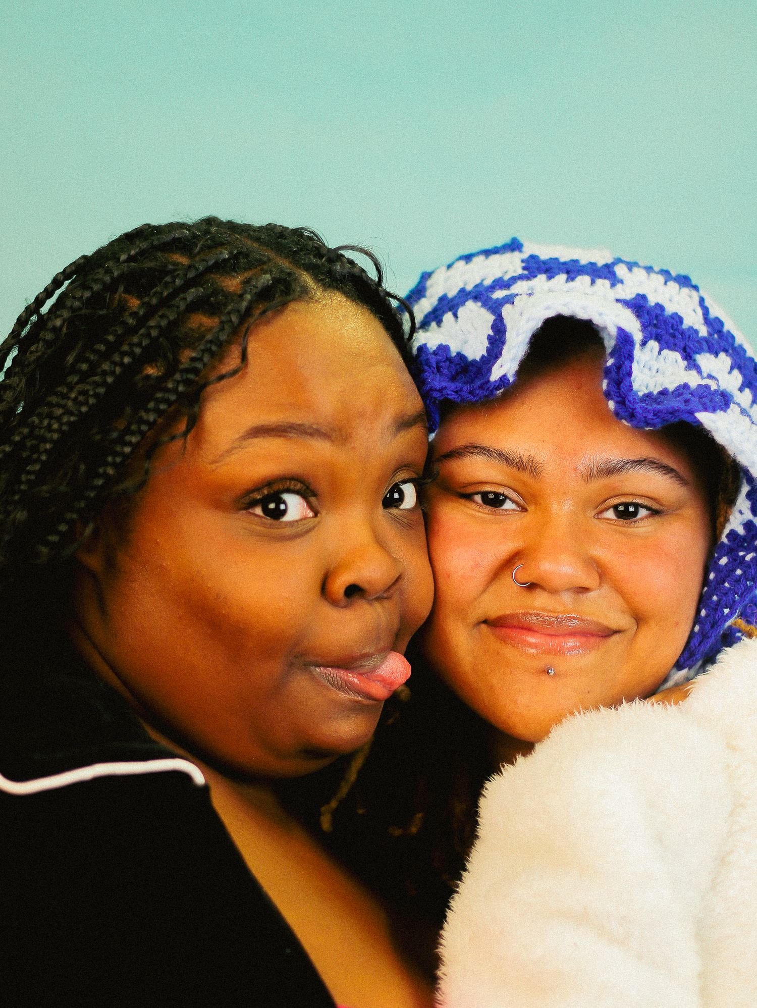 Two women pose cheek to cheek, one sticking out her tongue, against a light blue background.