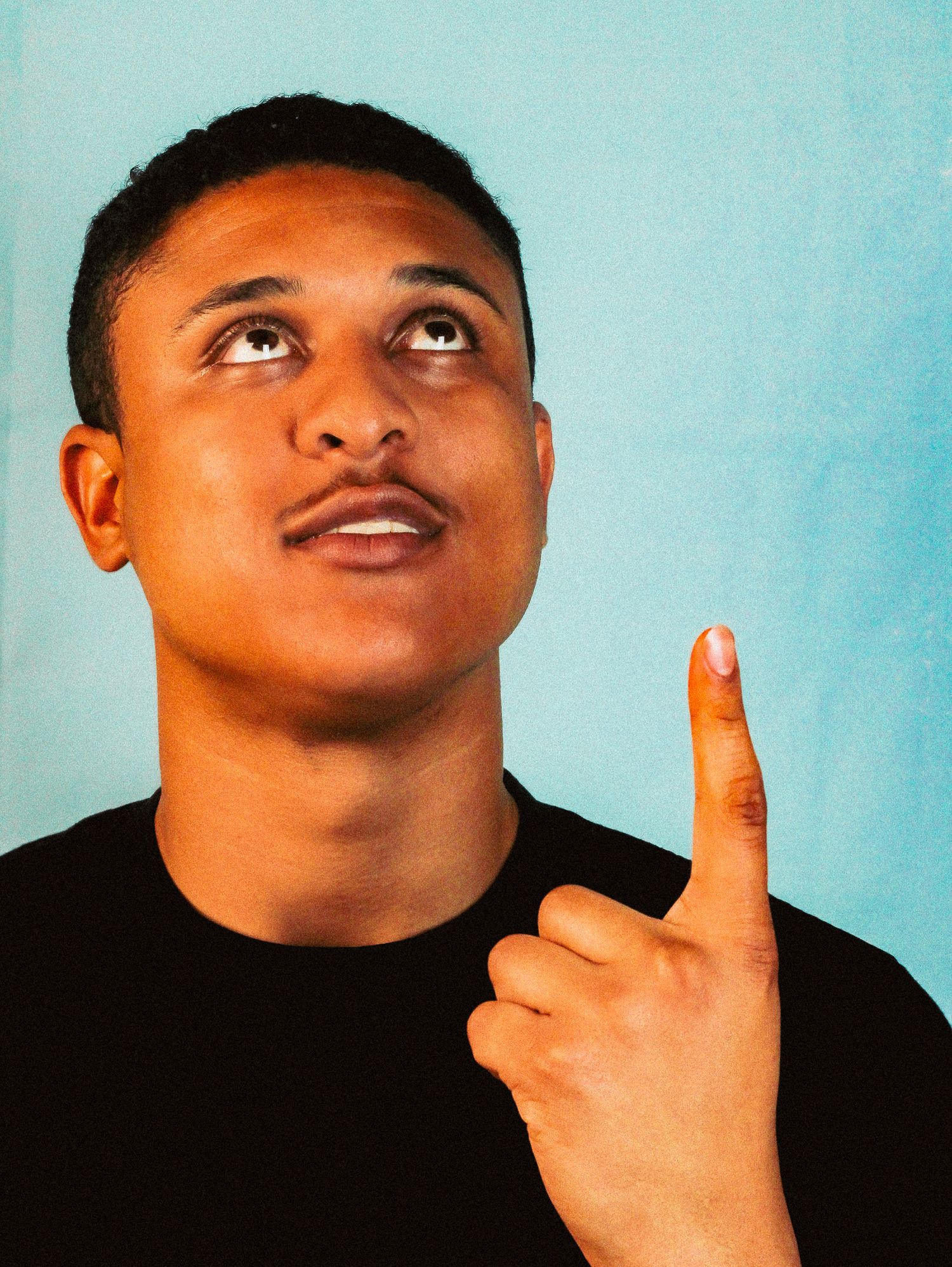 Young man pointing finger upward with a thoughtful expression on teal background.