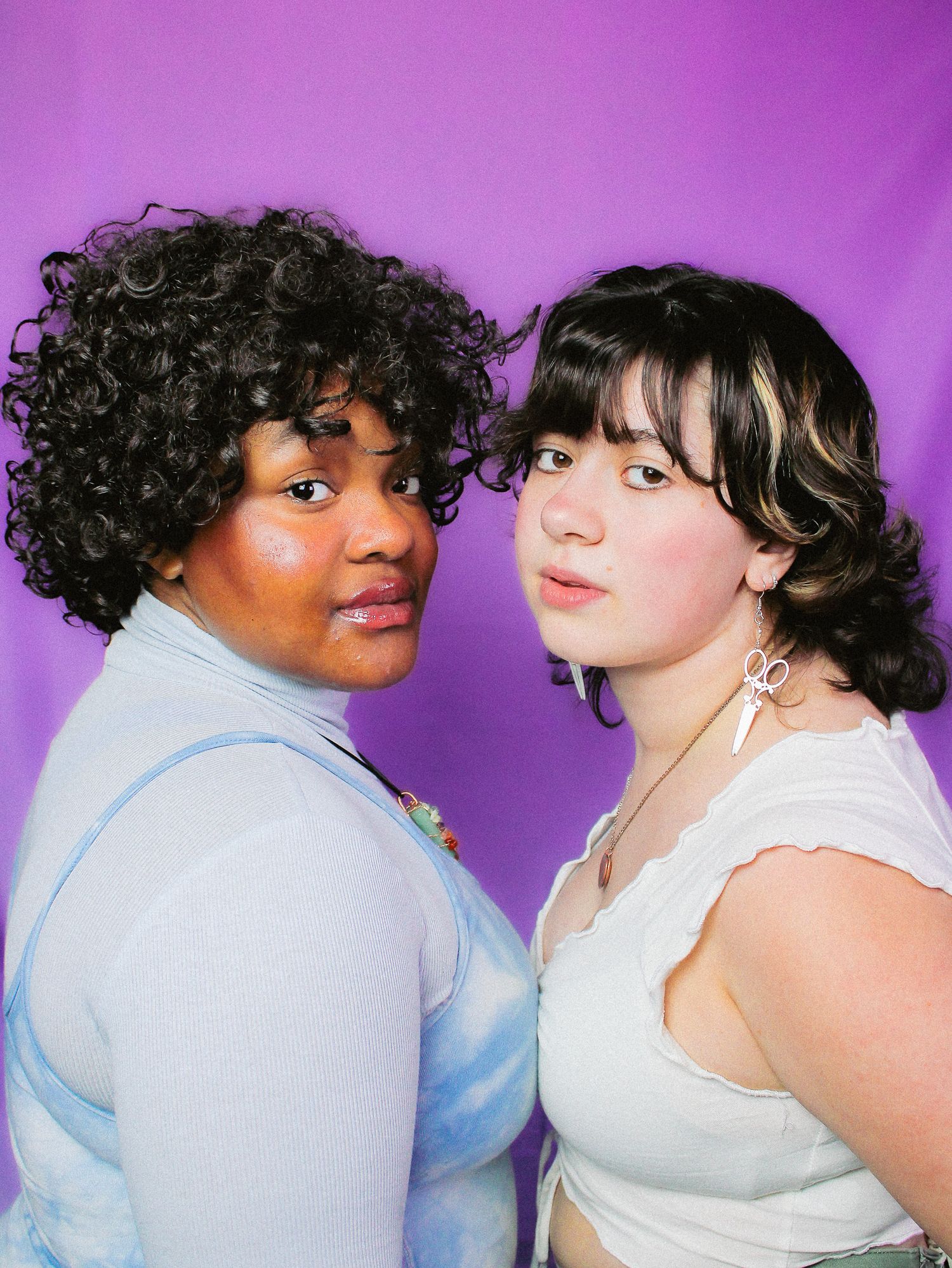Two young women stand close together against a purple backdrop, wearing casual tops and delicate jewelry.