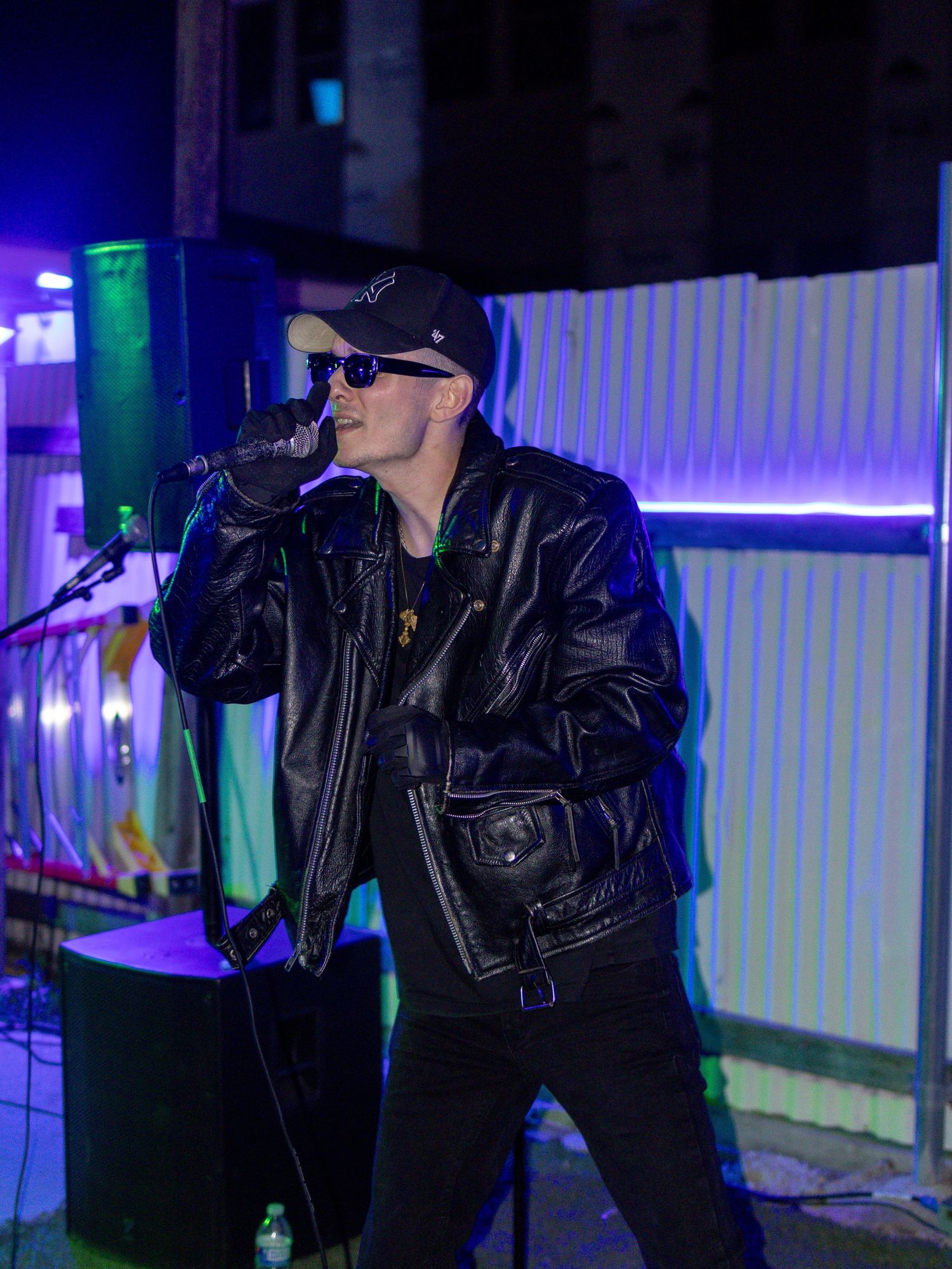 Singer in leather jacket and cap performs with microphone against illuminated backdrop.
