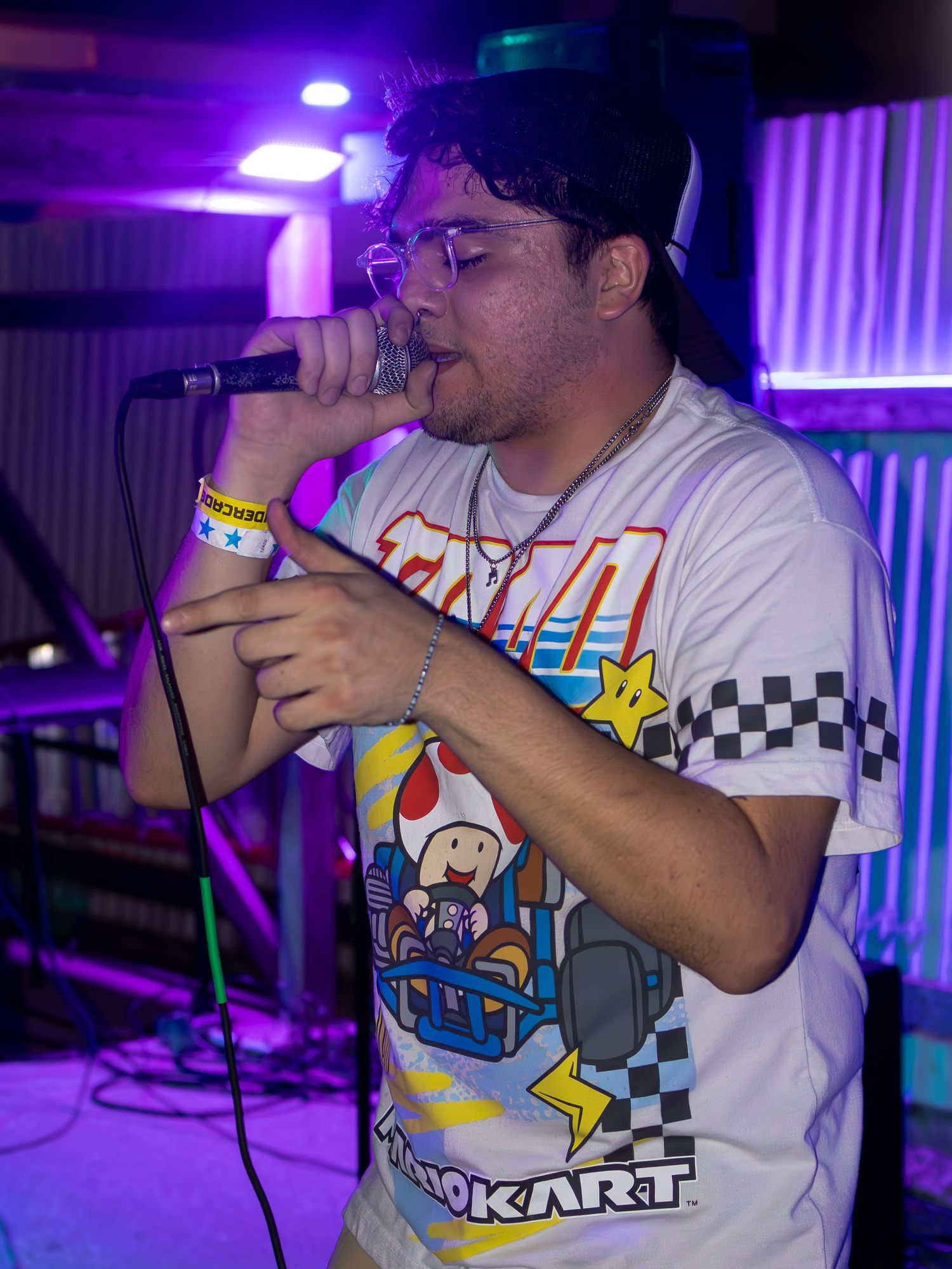 Side view of performer in Mario Kart shirt singing into microphone under purple stage lights.