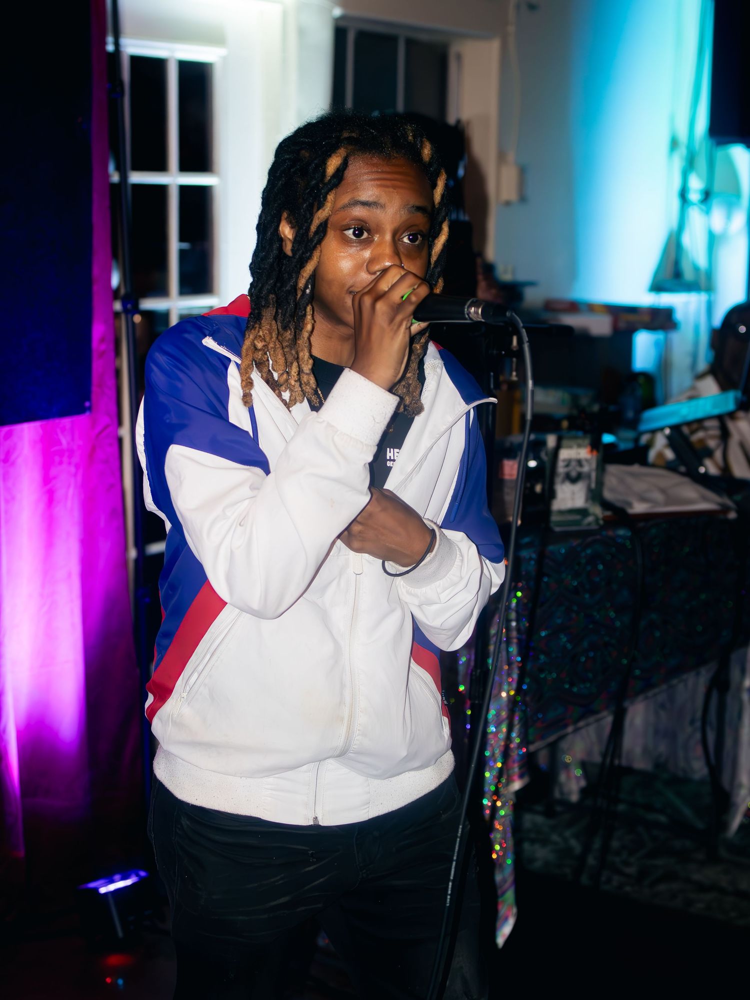 Performer in white and blue jacket singing passionately into microphone with colored lights in background.