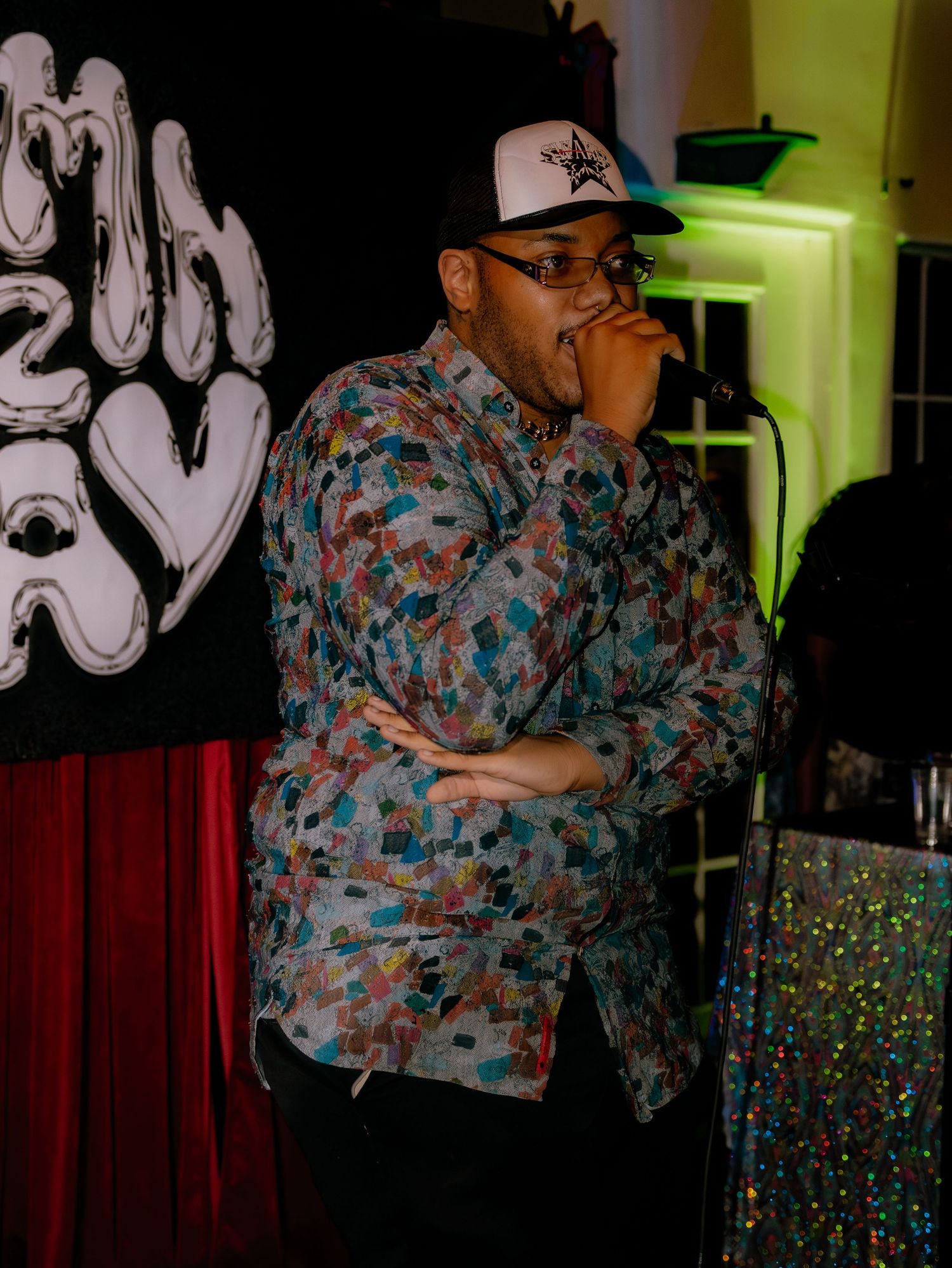 Stage performer in colorful outfit singing against neon-lit graffiti backdrop.