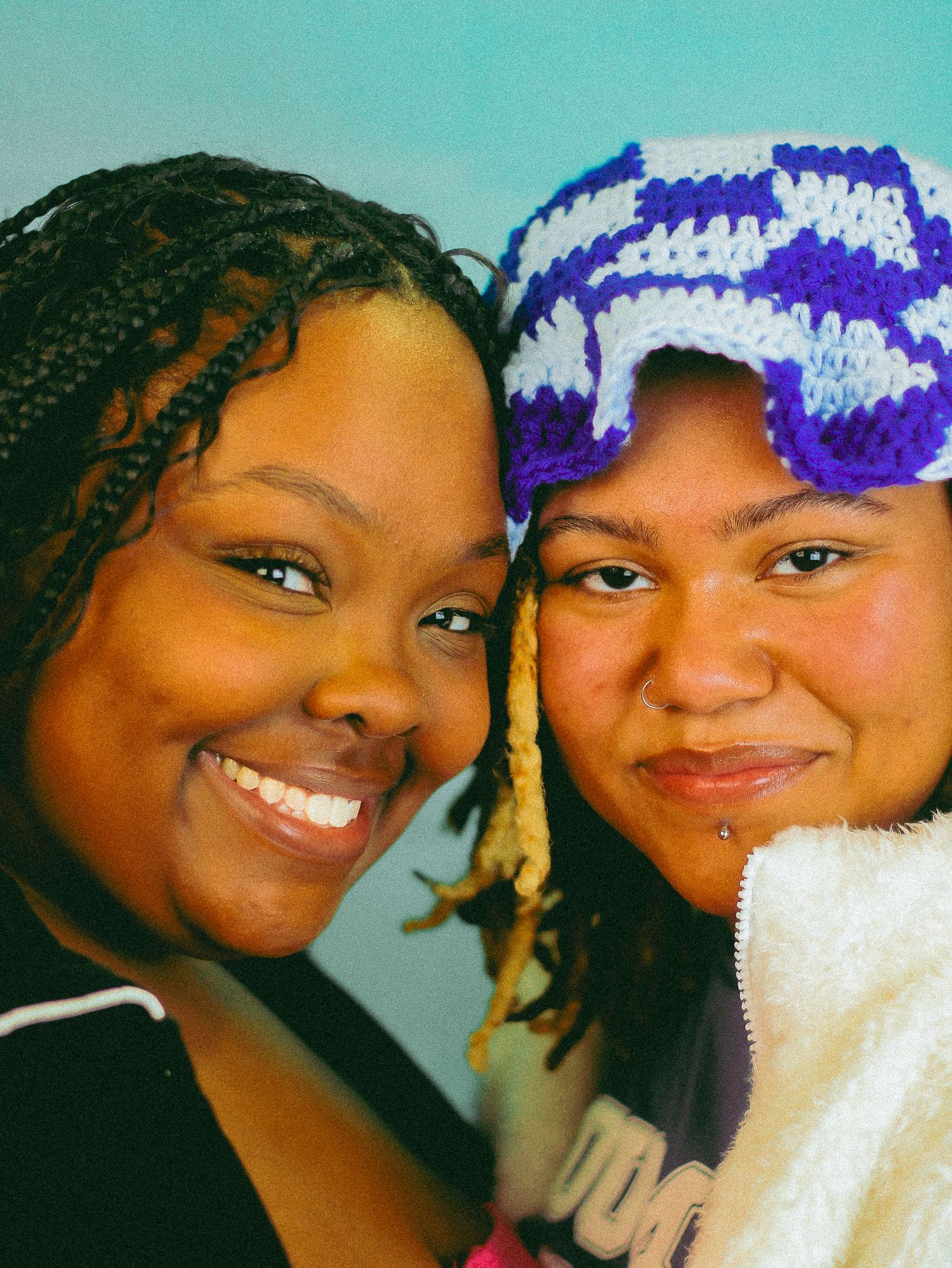 Two young women smiling closely together, one wearing a blue and white crocheted hat against a teal background.
